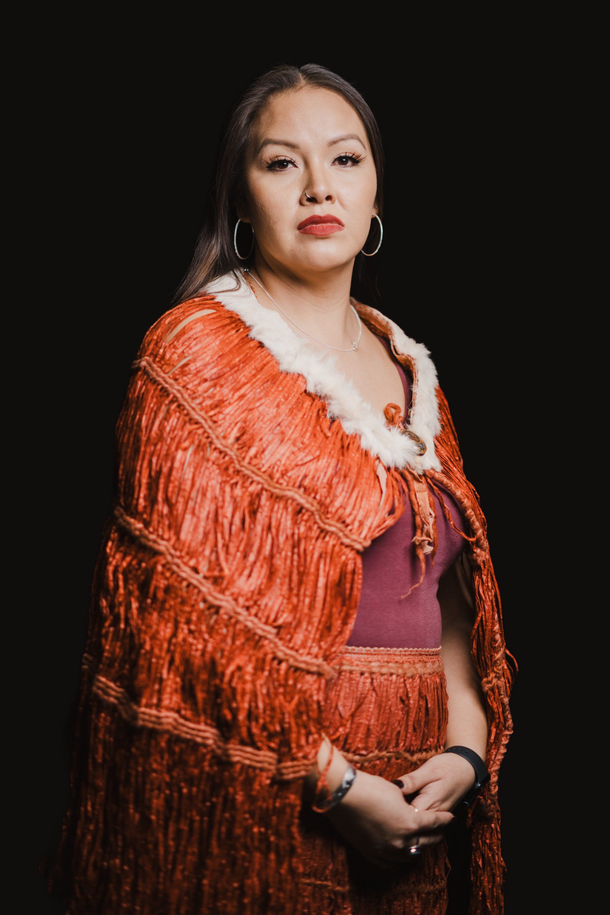 Woman in traditional orange cedar-bark regalia with white fur trim stands against black background, gazing thoughtfully aside