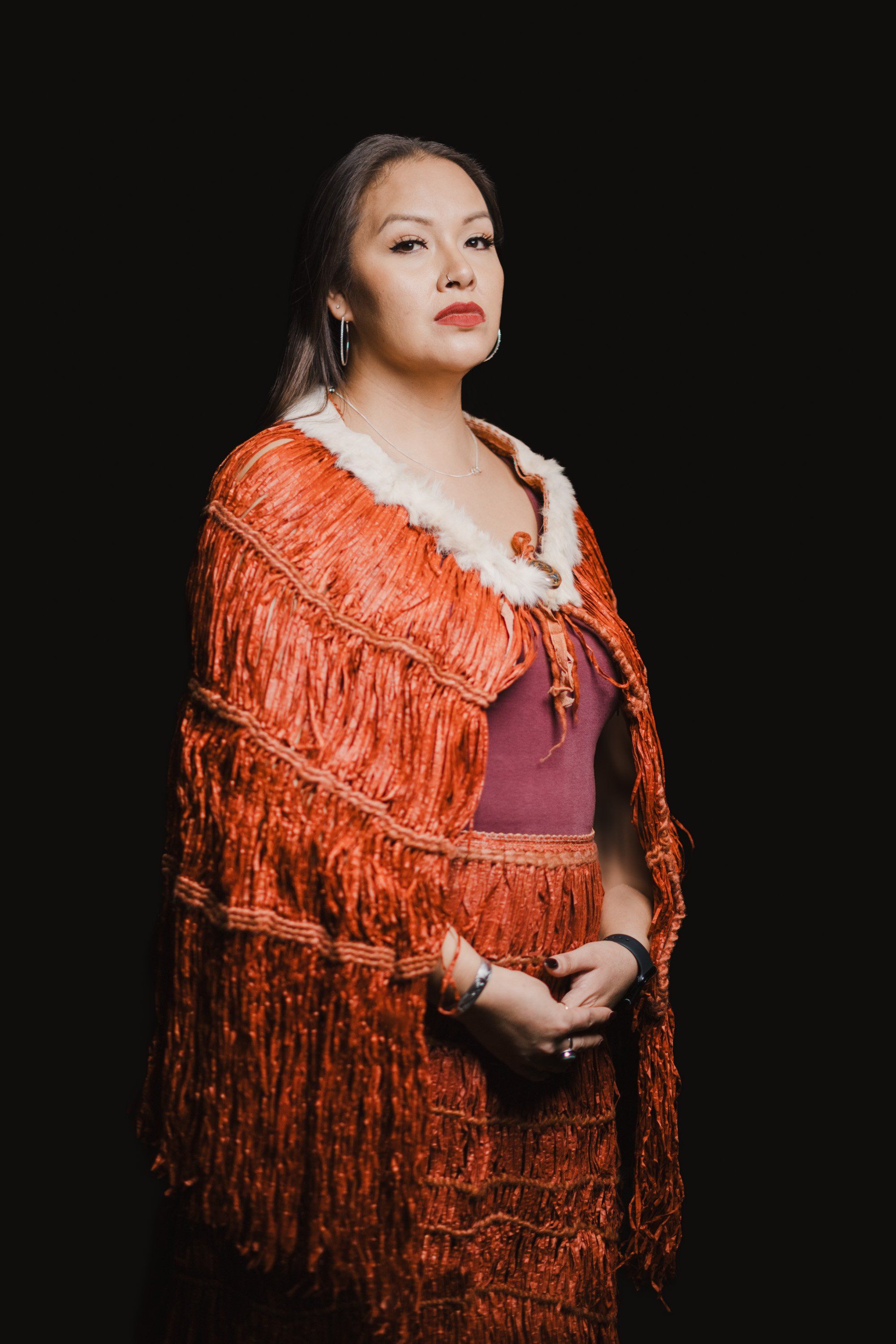 Woman in traditional Coast Salish regalia with orange fringed shawl and burgundy dress gazes thoughtfully against black background