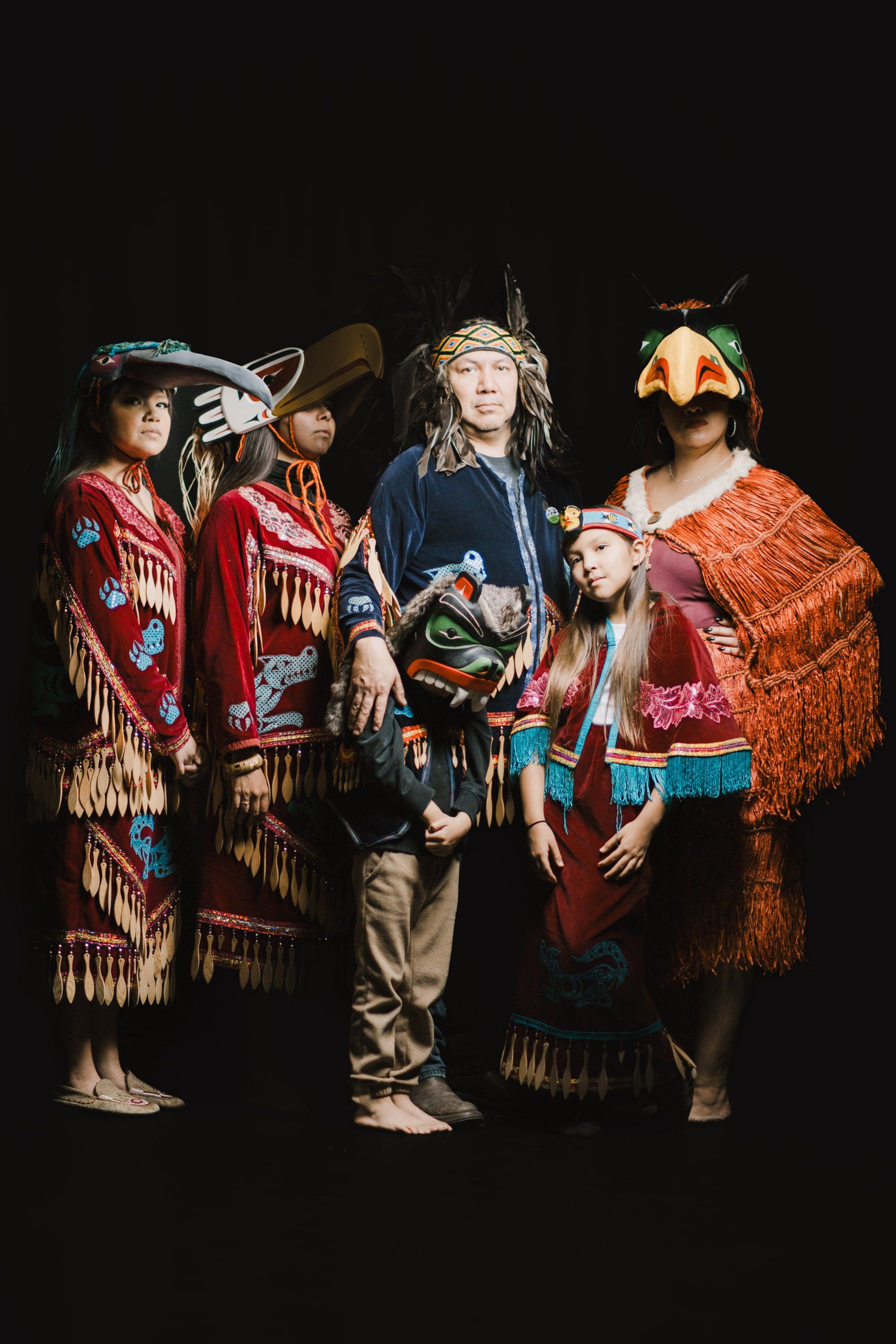 Four performers in elaborate traditional Coast Salish regalia stand against black, wearing carved masks and fringed blankets in rich reds and oranges