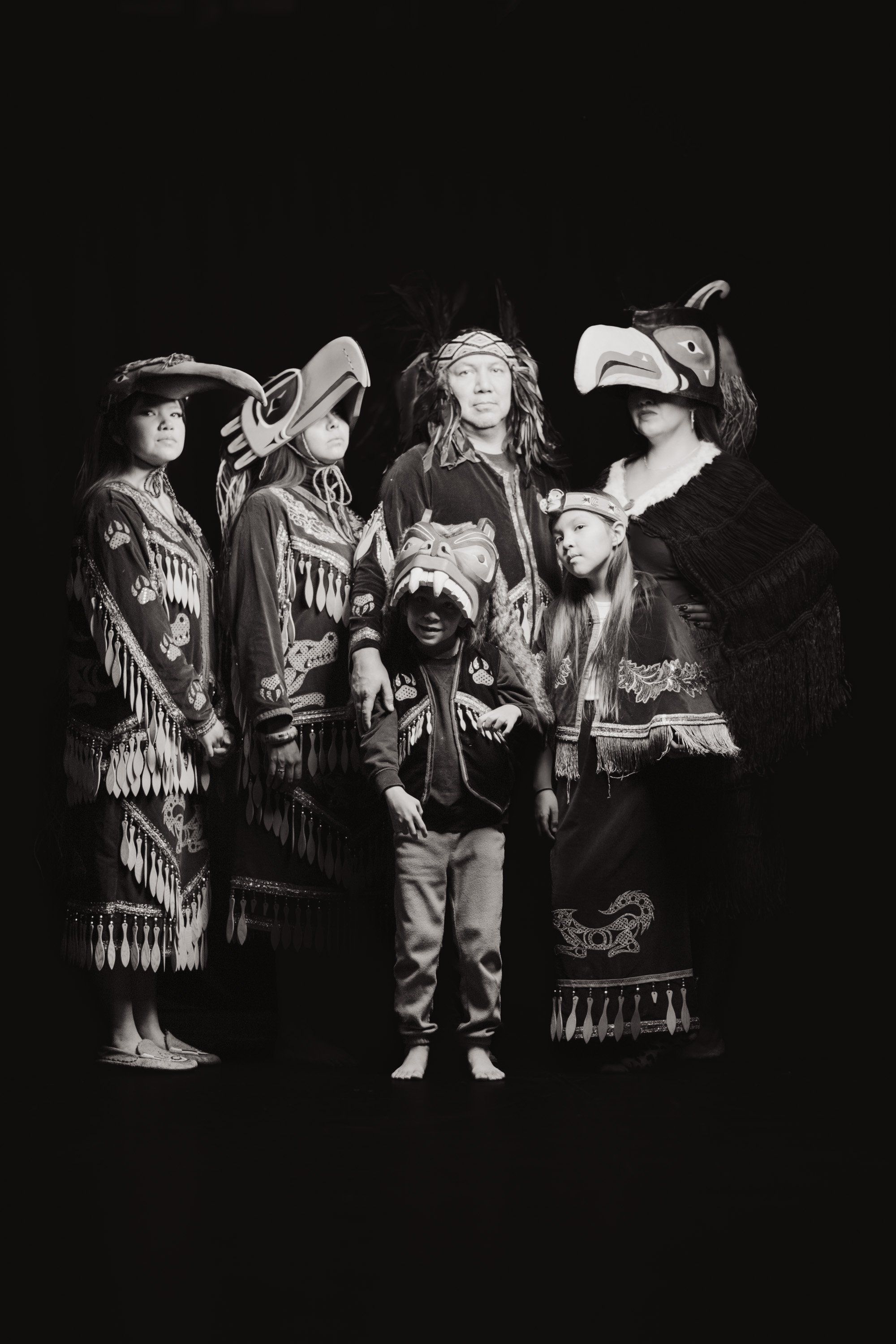 Four figures in elaborate regalia and traditional dress stand together against a dark background, illuminated in dramatic chiaroscuro lighting that highlight…