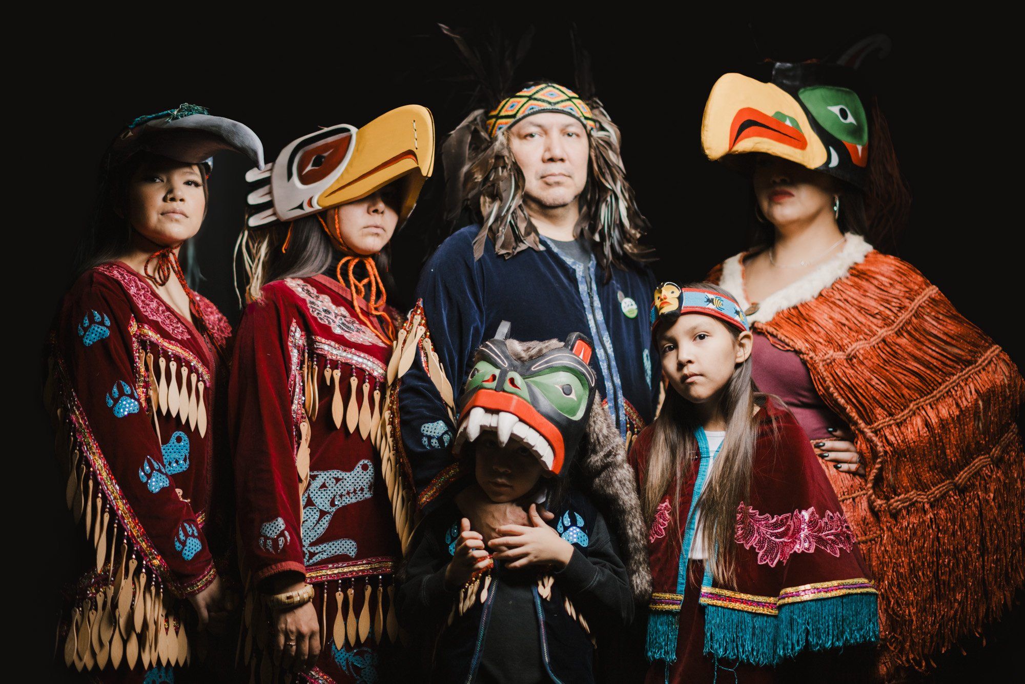 Five Coast Salish family members in vibrant traditional regalia with carved masks and fringed blankets stand together against a black background