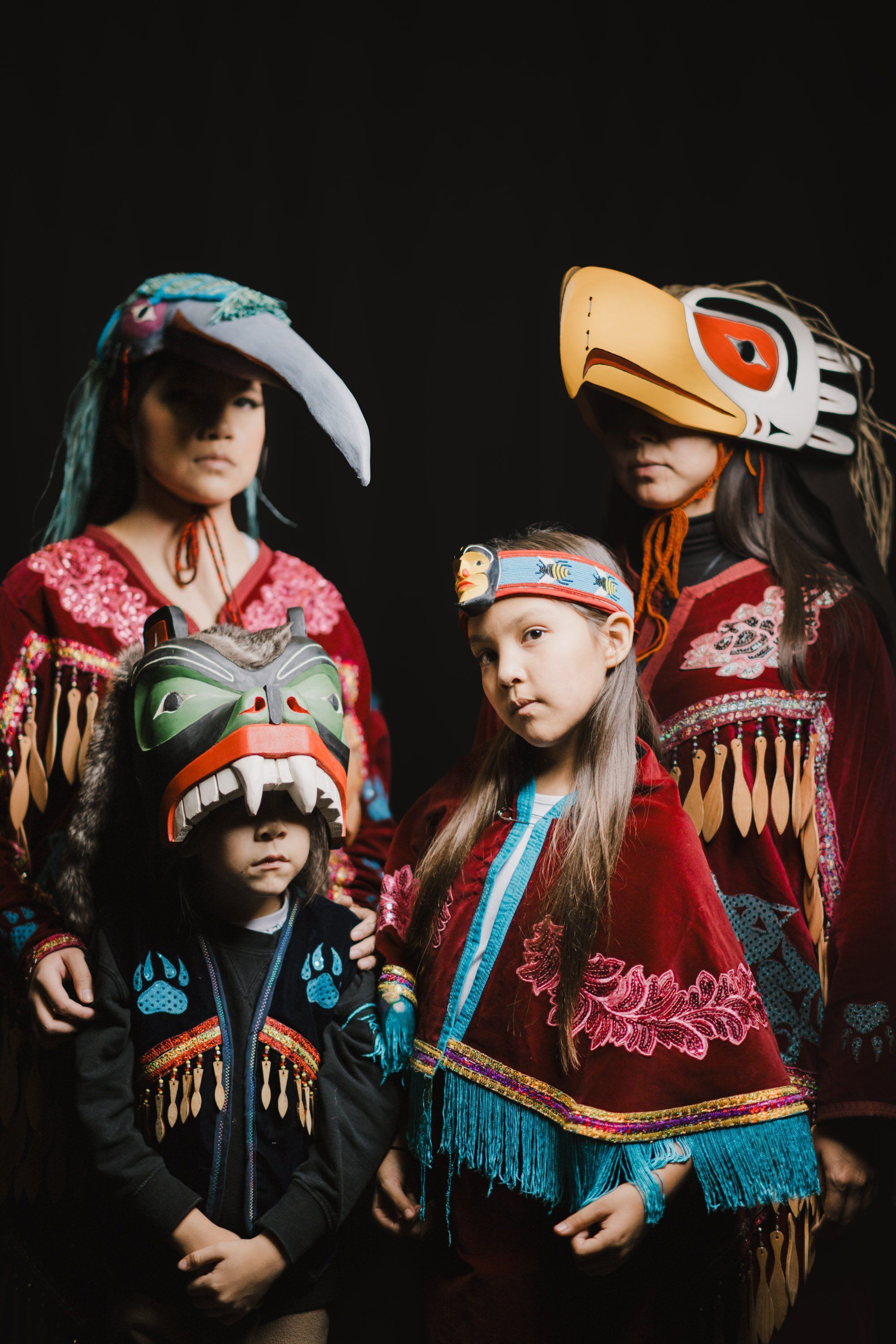 Four children in vibrant Coast Salish regalia with painted wooden masks and ornate button blankets pose together against a dark background