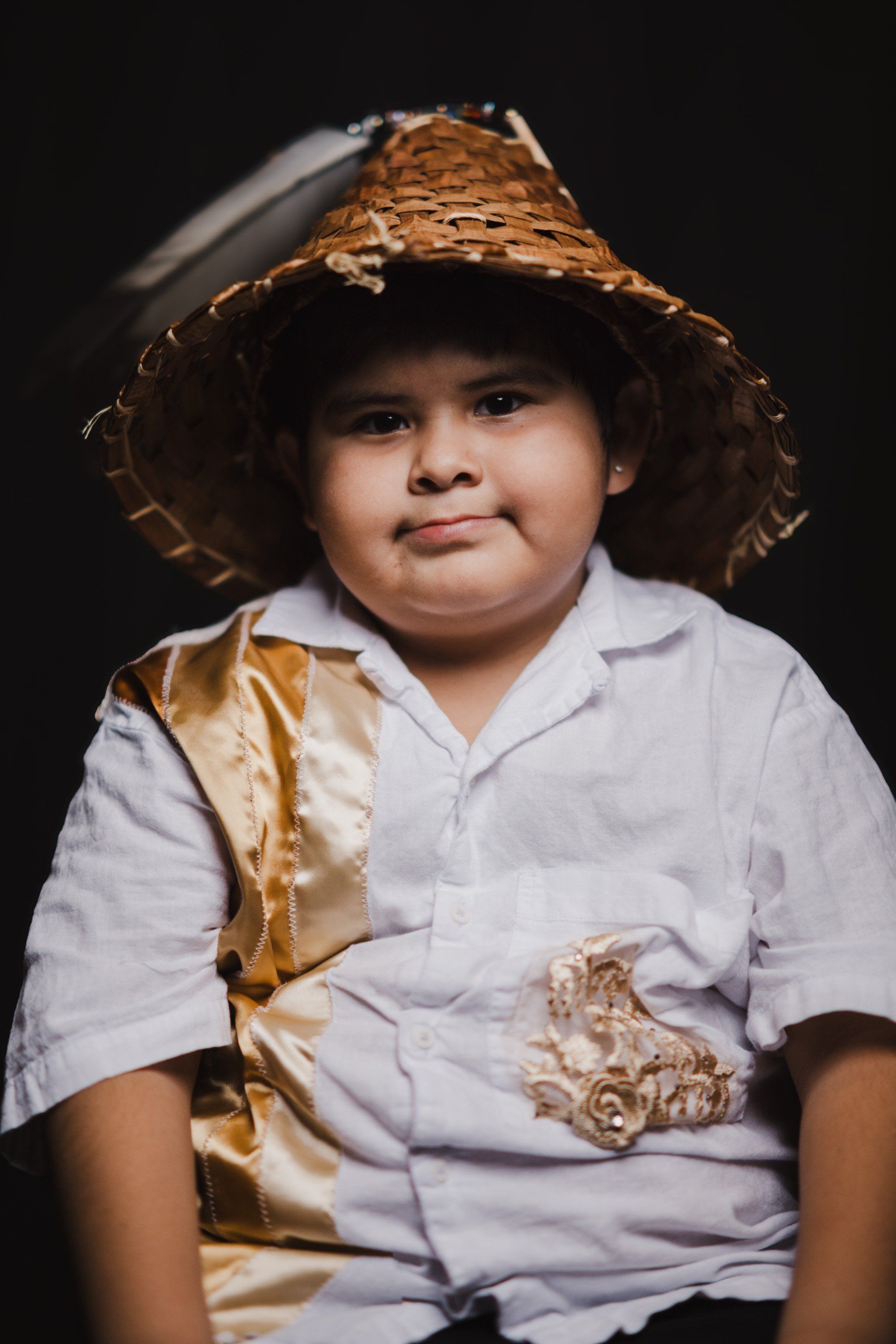 Young child in white shirt and golden sash wearing woven straw hat, gazing thoughtfully against dark background