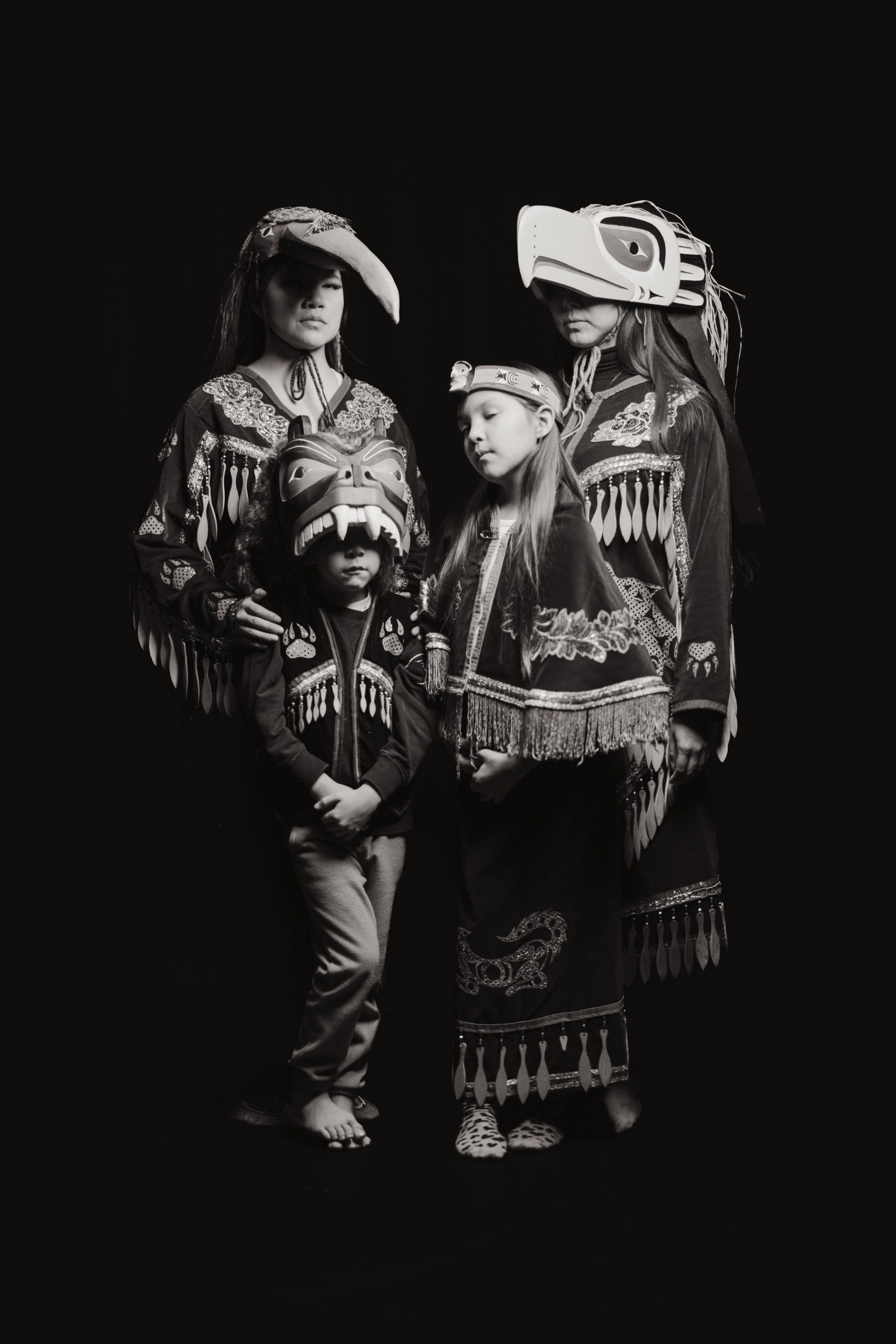 Three figures in traditional Coast Salish regalia with fringe and formline design stand together against black, illuminated by dramatic side lighting