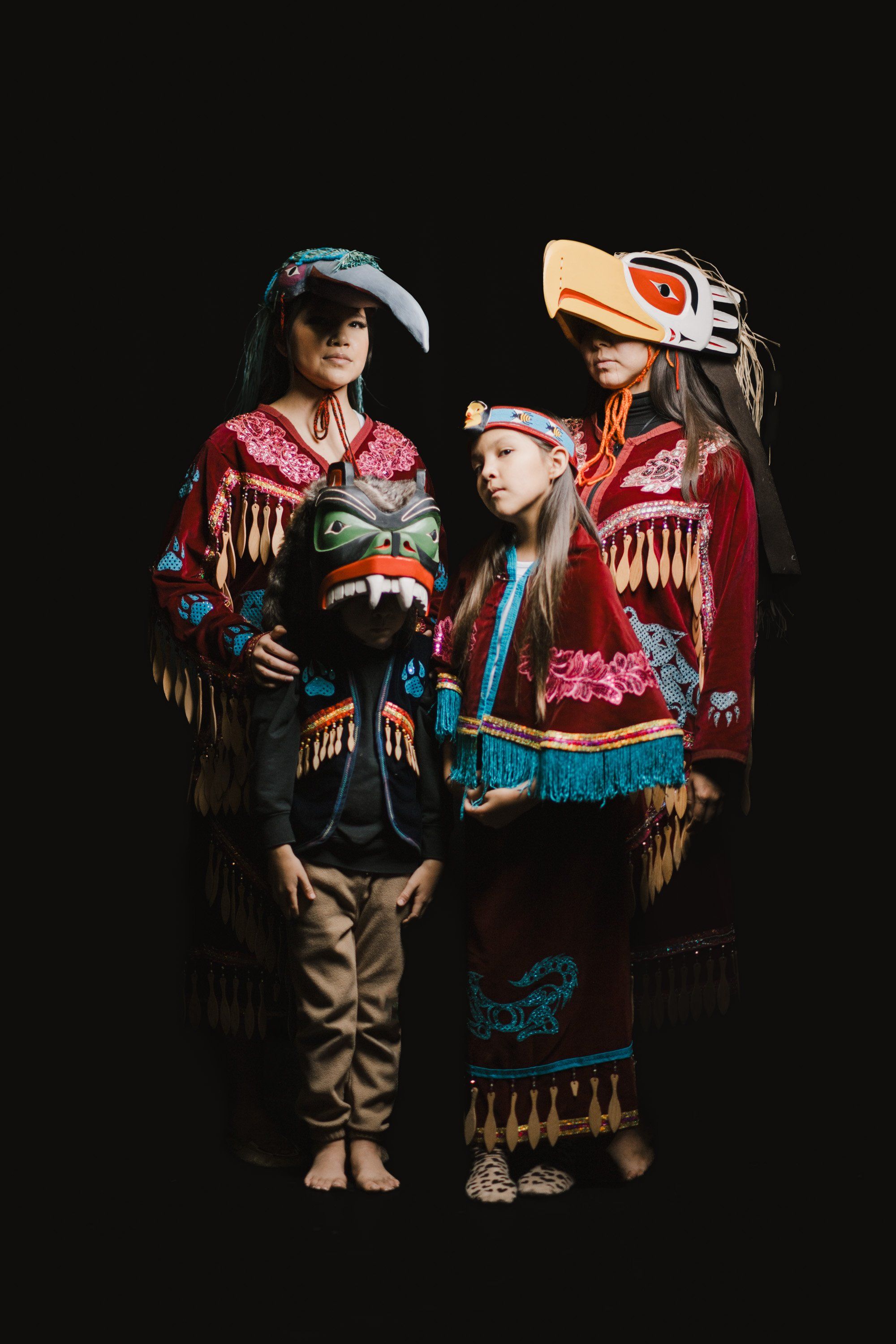 Three Coast Salish family members in vibrant regalia with red, blue, and gold beadwork stand together against a black background, their traditional button bl…