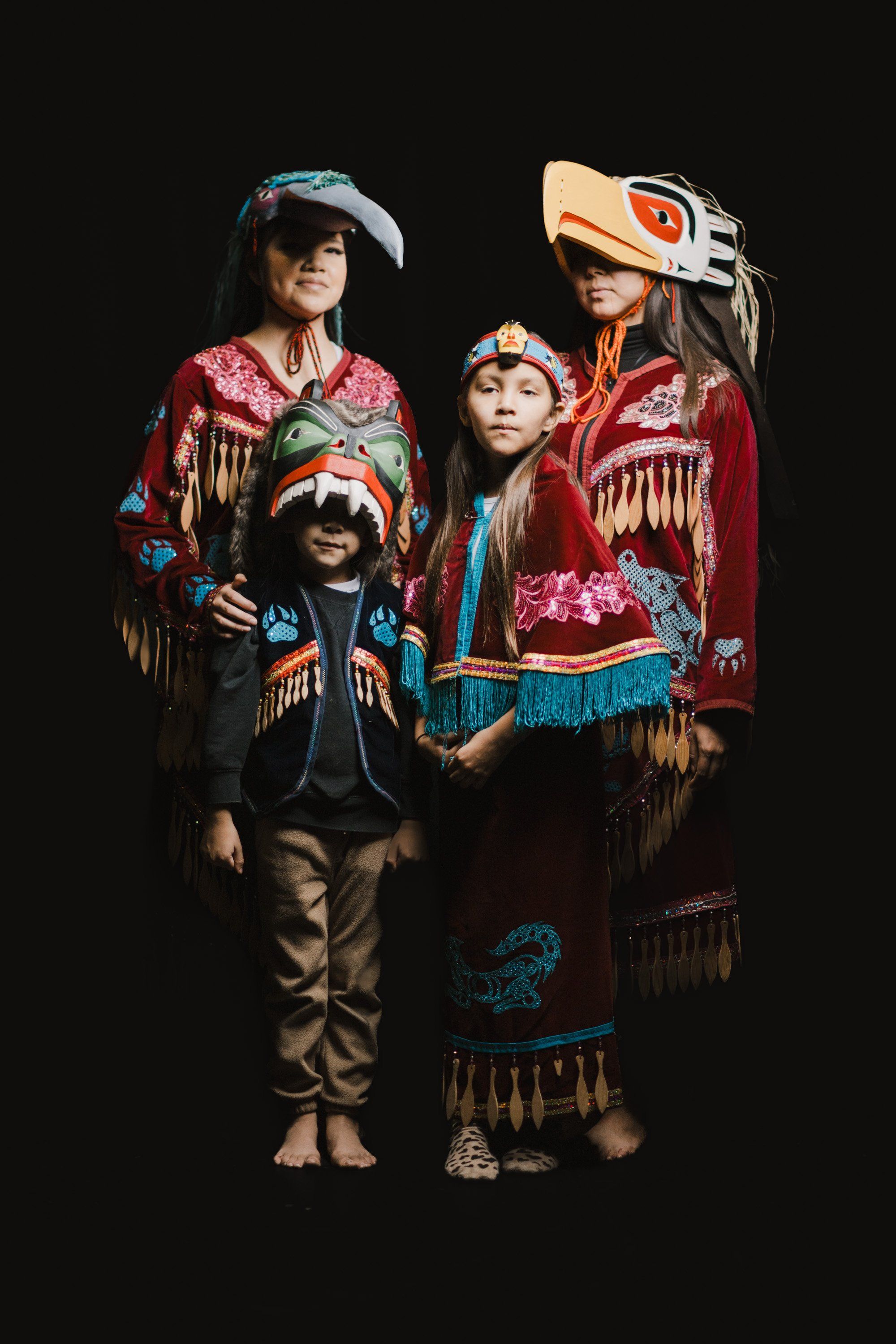 Three children in vibrant red and black regalia with fringe and formline designs pose together against a dark background, their ceremonial clothing and carve…