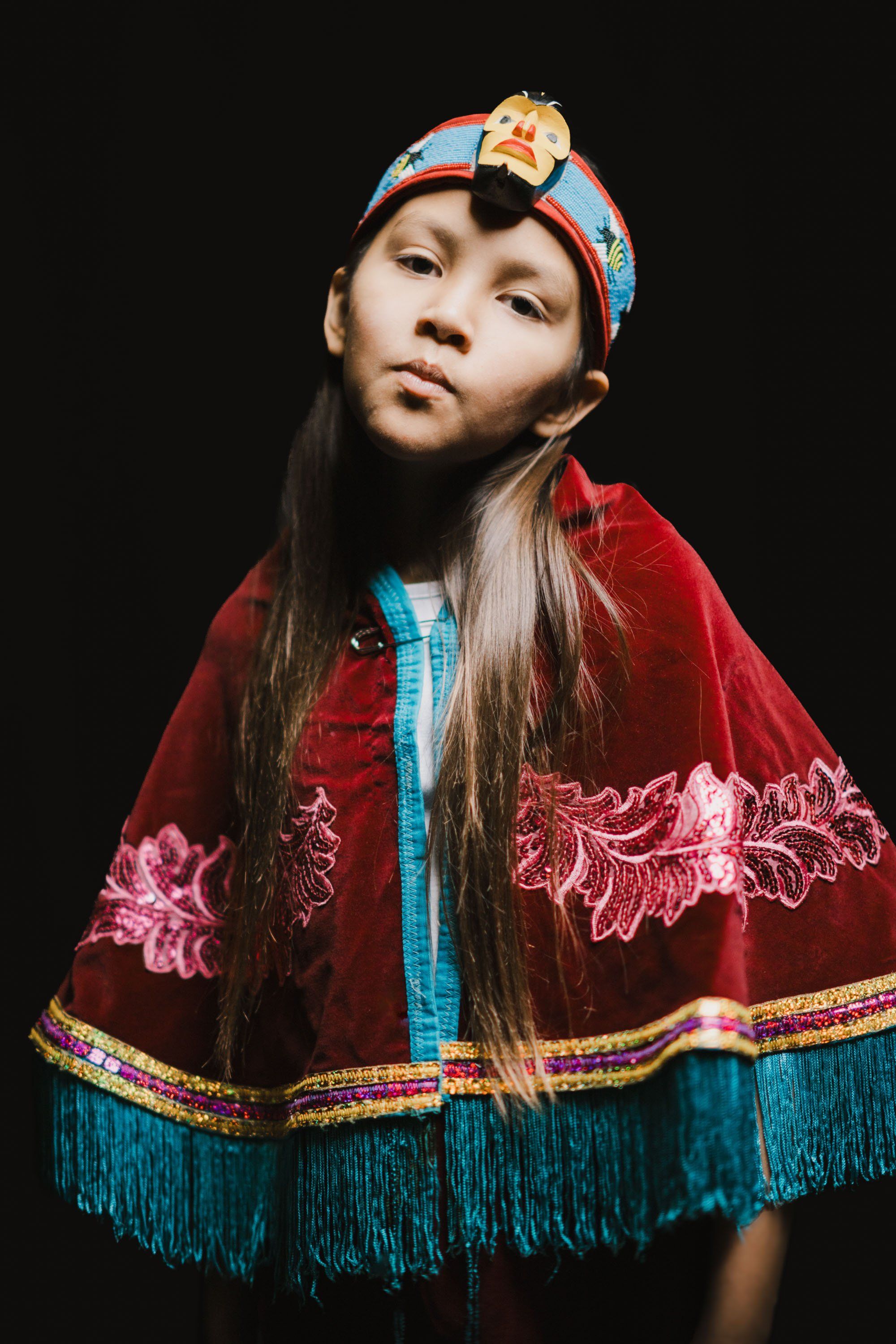 Young dancer in traditional Coast Salish regalia with red velvet cape and blue trim against black background, looking thoughtfully upward