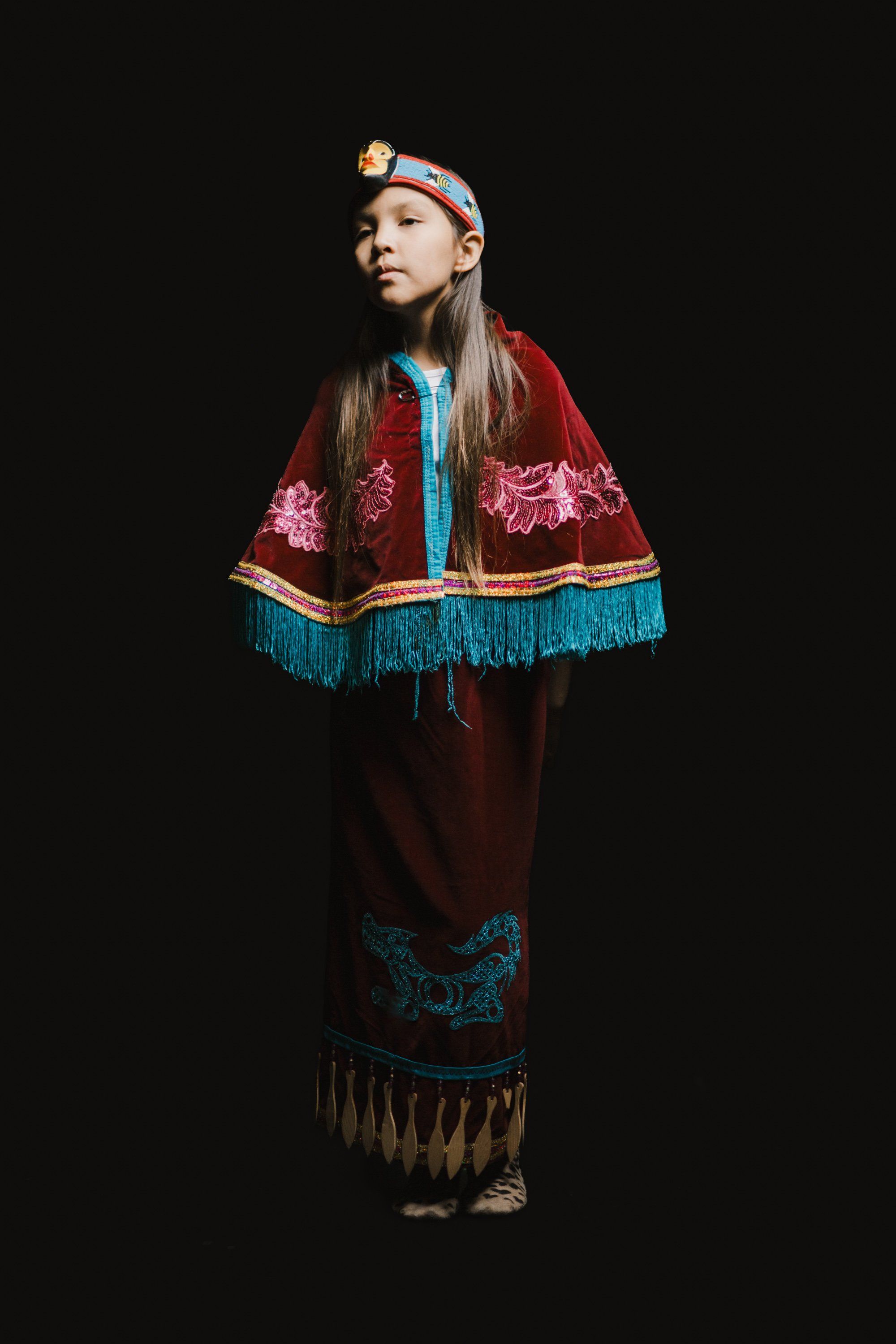 Young dancer in traditional Coast Salish regalia with burgundy button blanket and turquoise fringe against black background