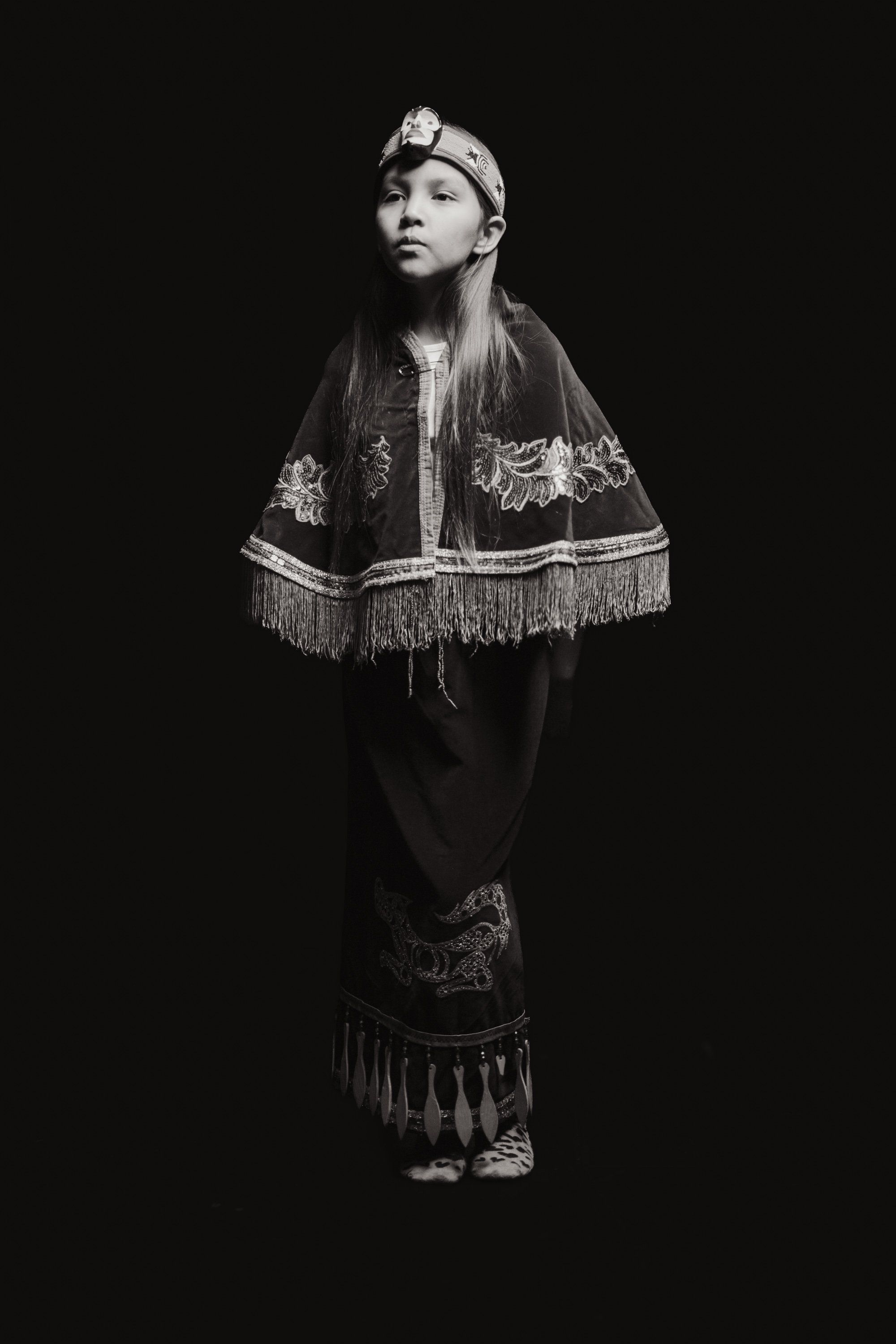 Woman in traditional Coast Salish regalia with embroidered cape and headpiece, gazing upward against black background
