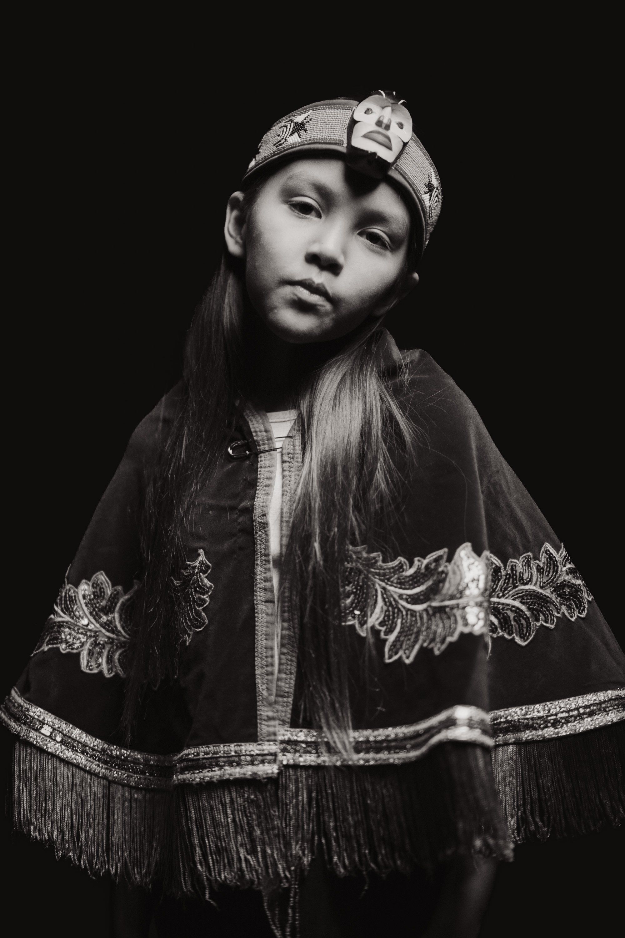 Young dancer in traditional Coast Salish regalia with fringed shawl and decorated cap, gazing downward against black background