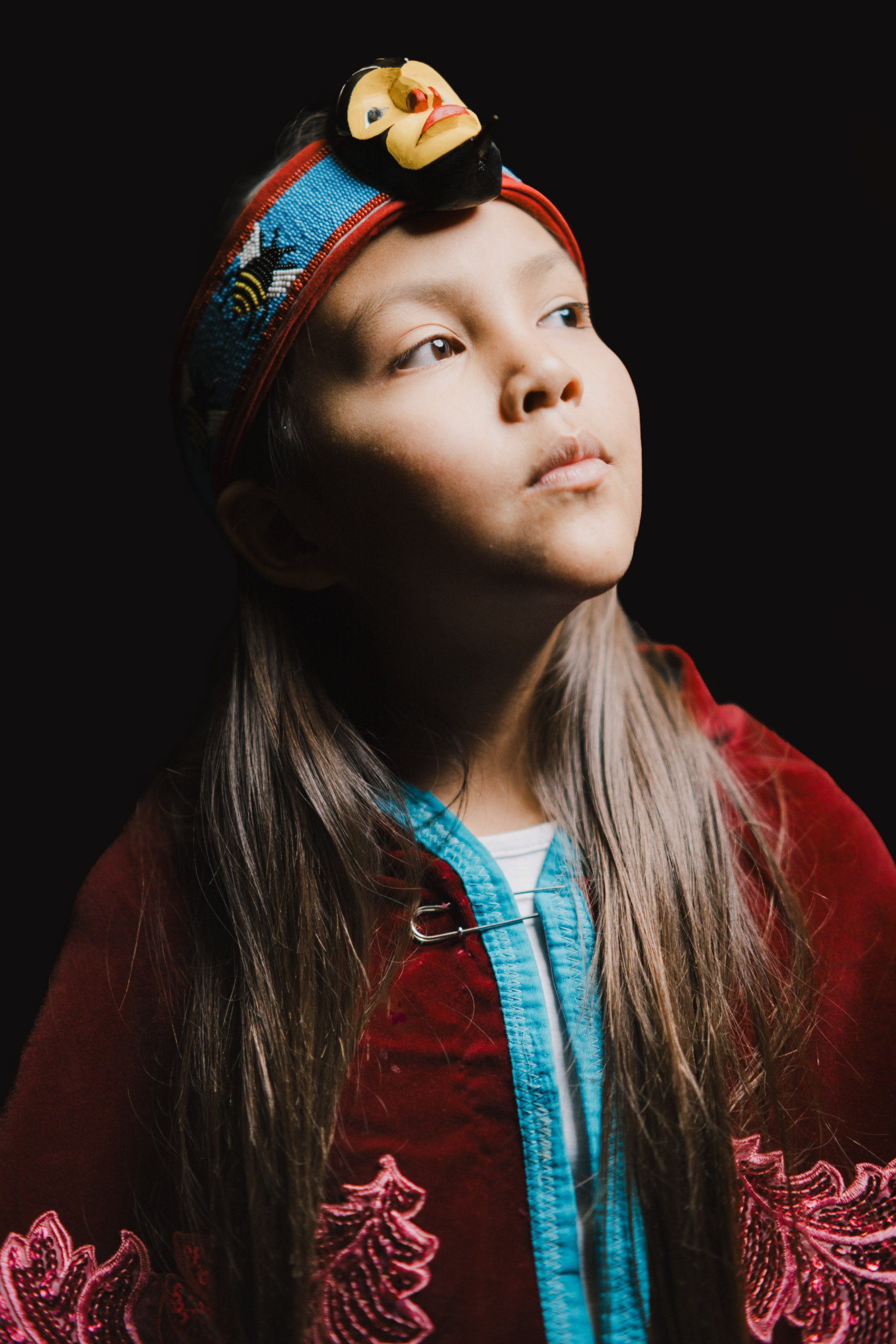 Young person in red embroidered jacket and turquoise shirt gazes upward, wearing ornate wooden and brass headdress against dark background