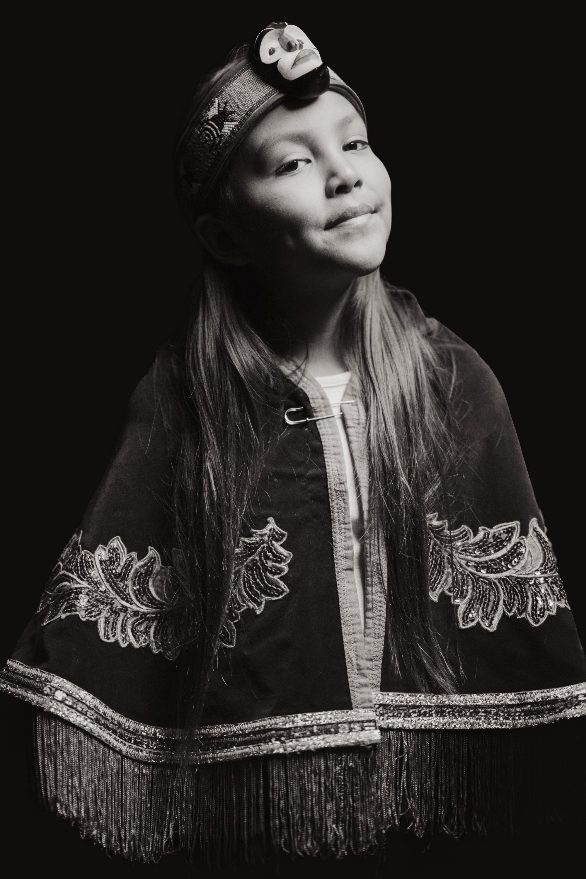Young person in traditional Coast Salish regalia with embroidered cape and hair ornament, gazing upward against dark background