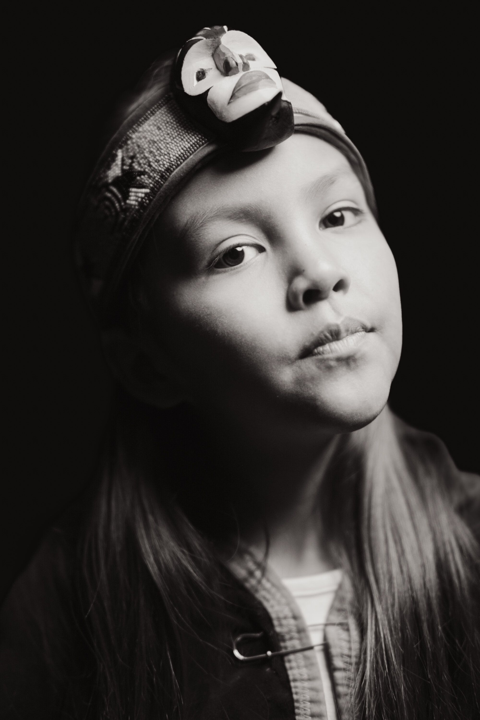 Young Coast Salish performer in dramatic black-and-white portrait, gazing upward with traditional ornament in hair against dark background