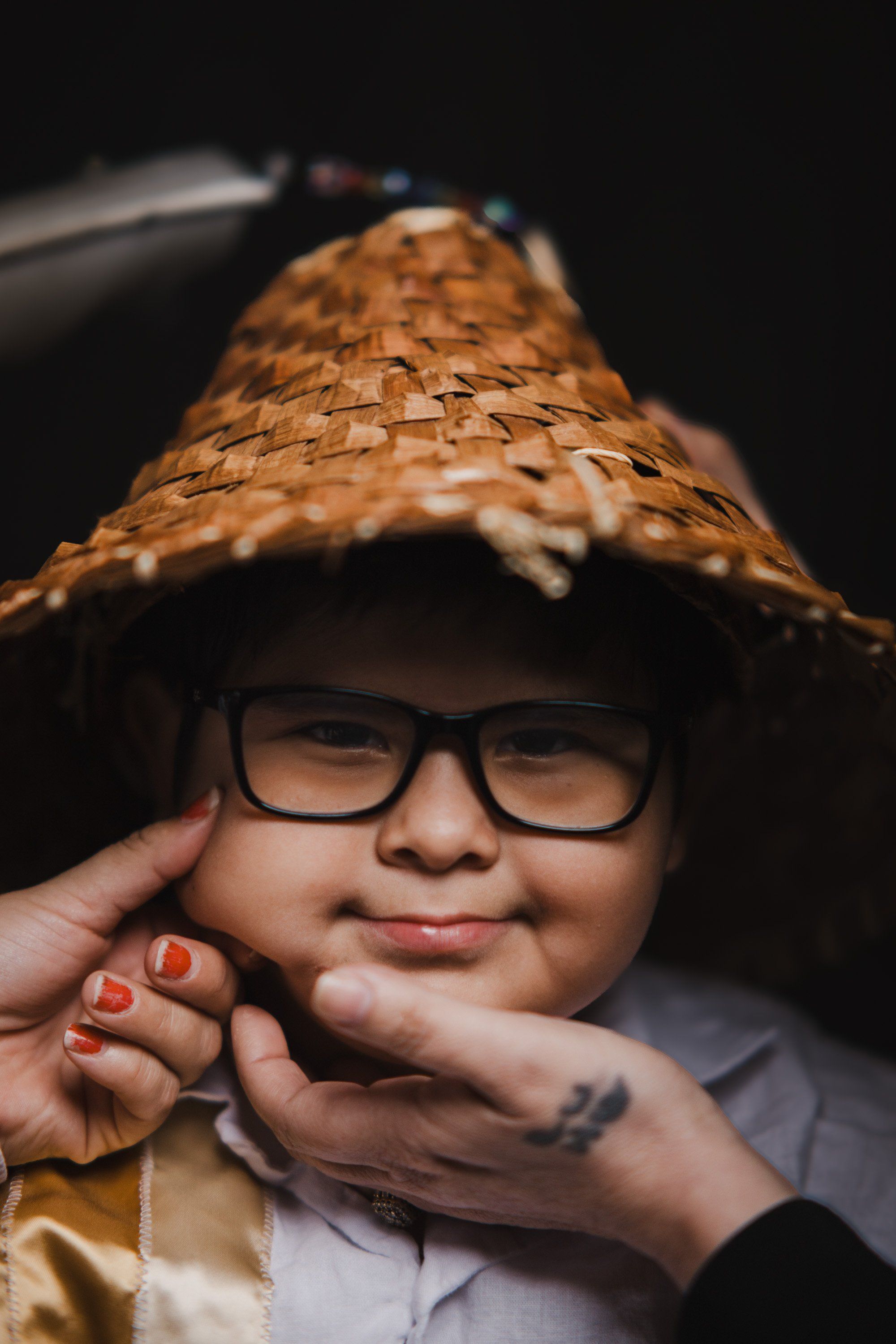 Young child wearing oversized straw hat and glasses, smiling gently while held by an adult's hands against a dark background