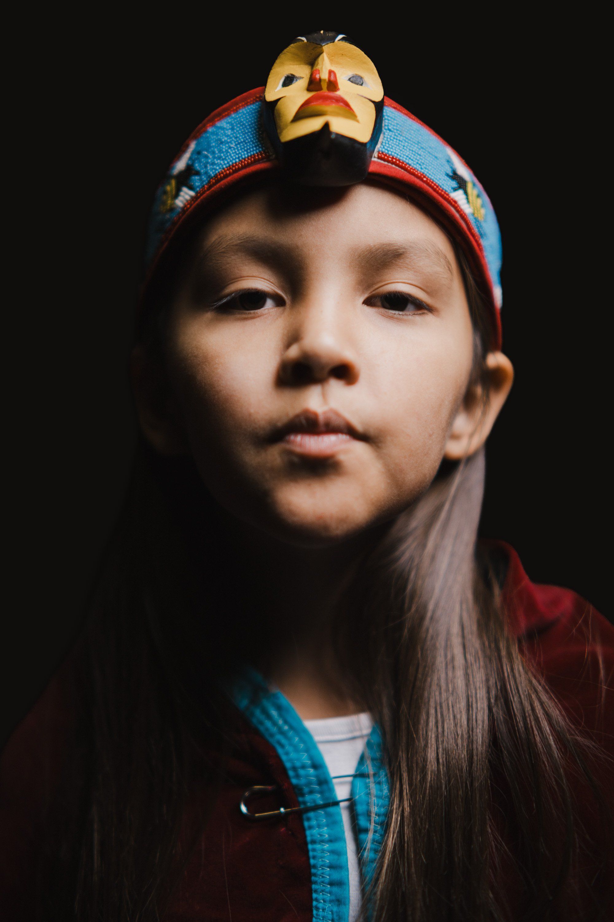 Young person wearing traditional Coast Salish regalia with blue and red woven headpiece and gold mask, against dark background