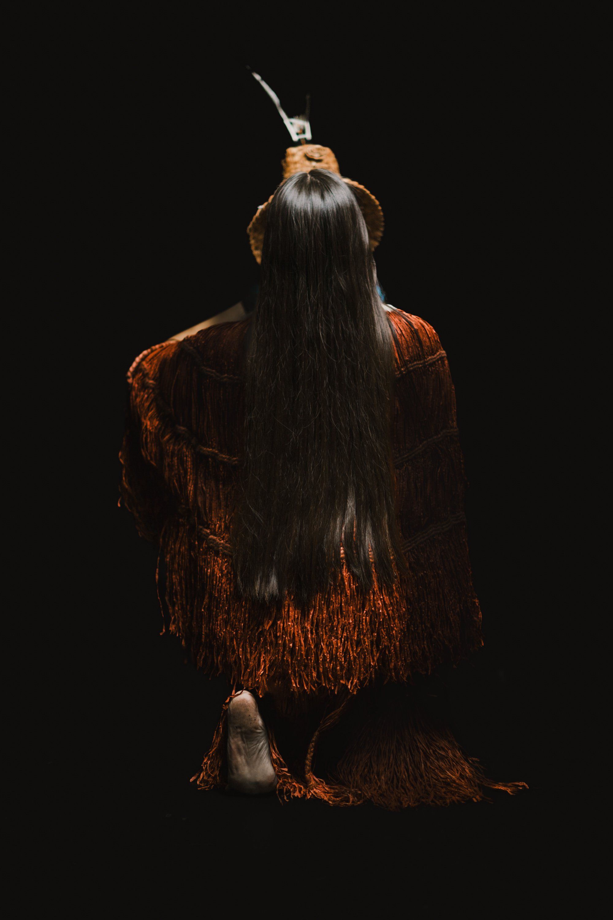 Person from behind wearing rust-colored cedar bark regalia and traditional headdress against black background, long dark hair flowing