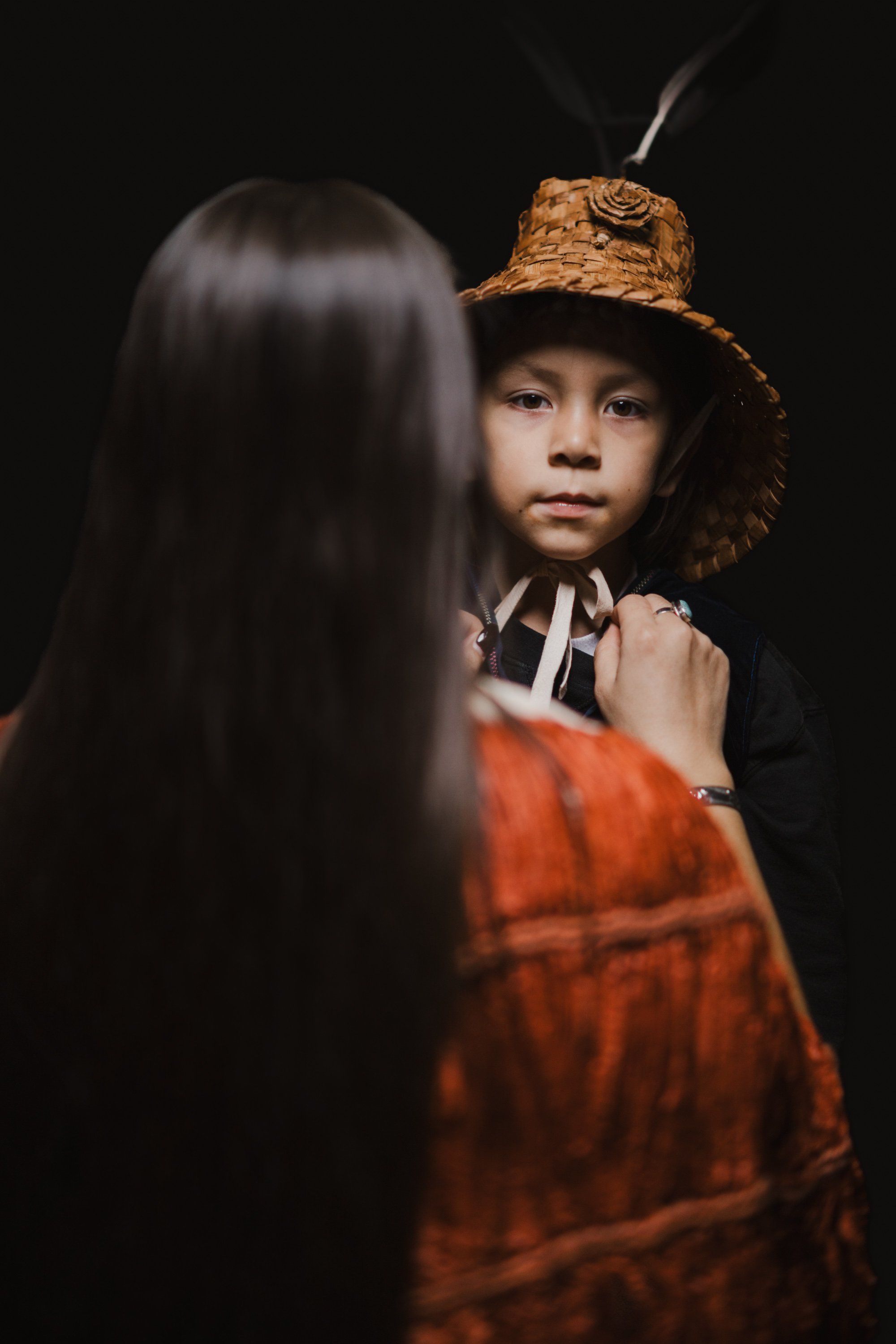 Child in straw hat and red regalia faces away from camera toward another person with long dark hair against black background