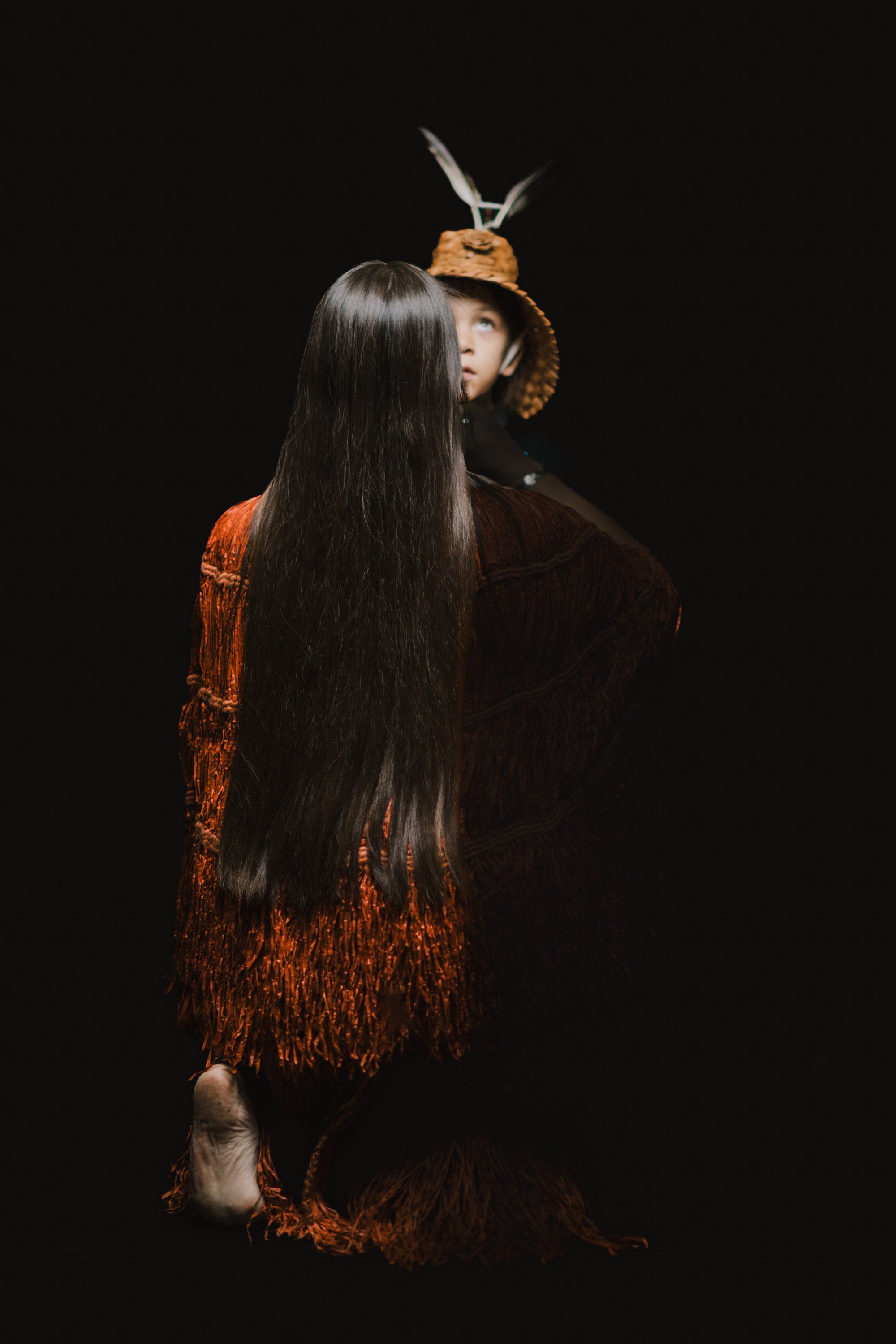 Woman with long black hair viewed from behind, wearing rust-orange traditional regalia, facing a figure in carved wooden mask and headdress against black bac…