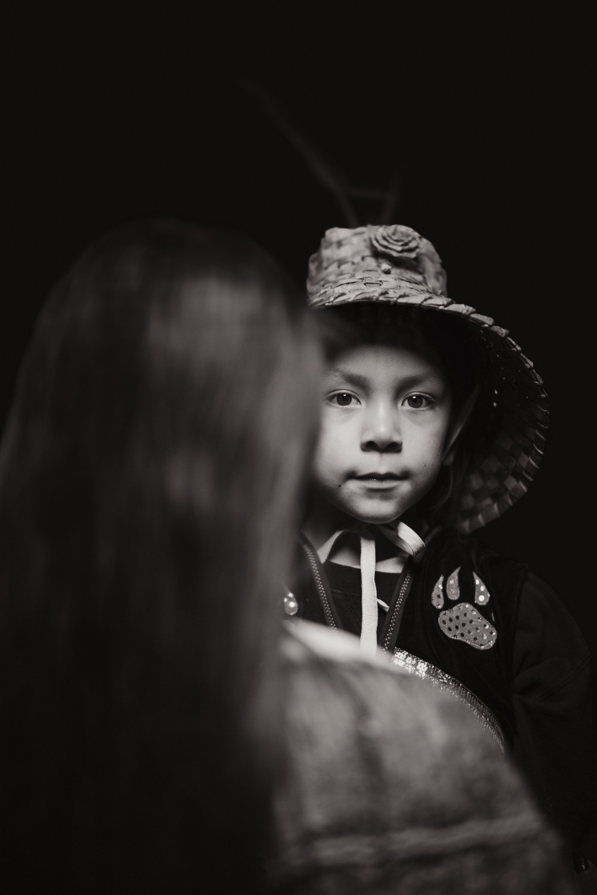 Young child in traditional regalia and woven hat faces camera with solemn expression, backlit against dark background with blurred figure beside them