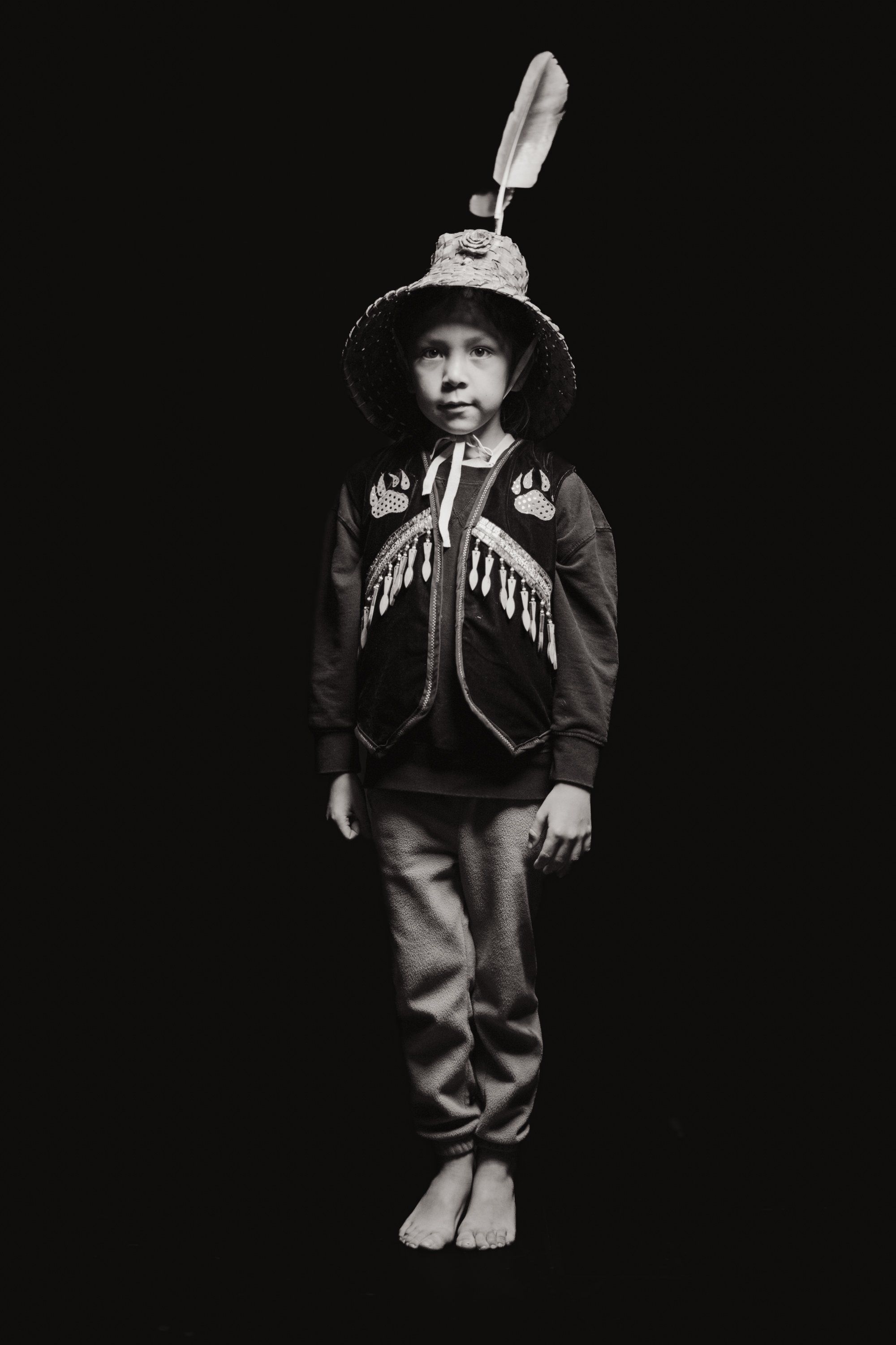 Young child wearing traditional Coast Salish regalia with fringed vest and decorative hat topped with feather, standing barefoot against black background