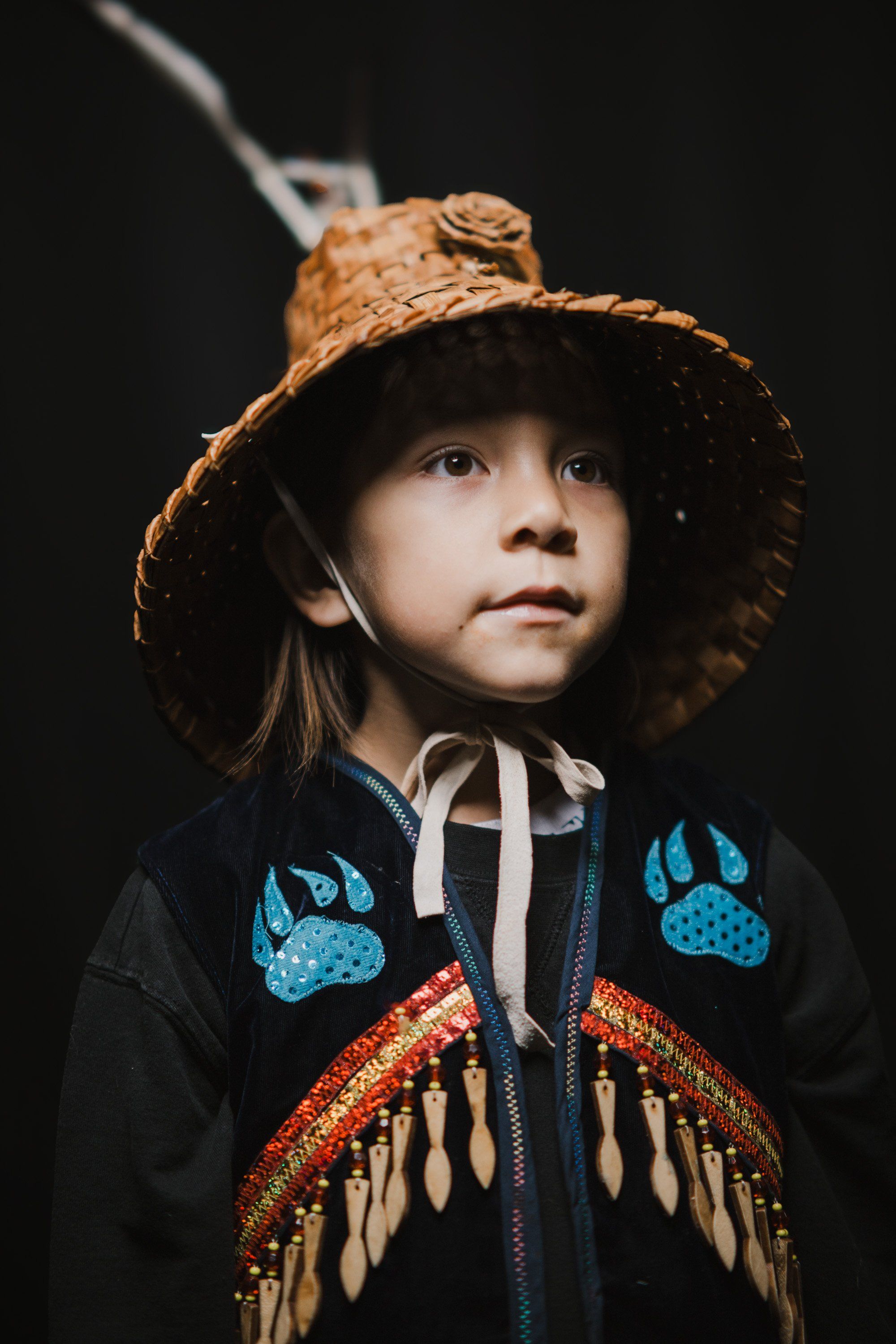Child in straw hat and ornate beaded regalia with fringe gazes upward against dark background