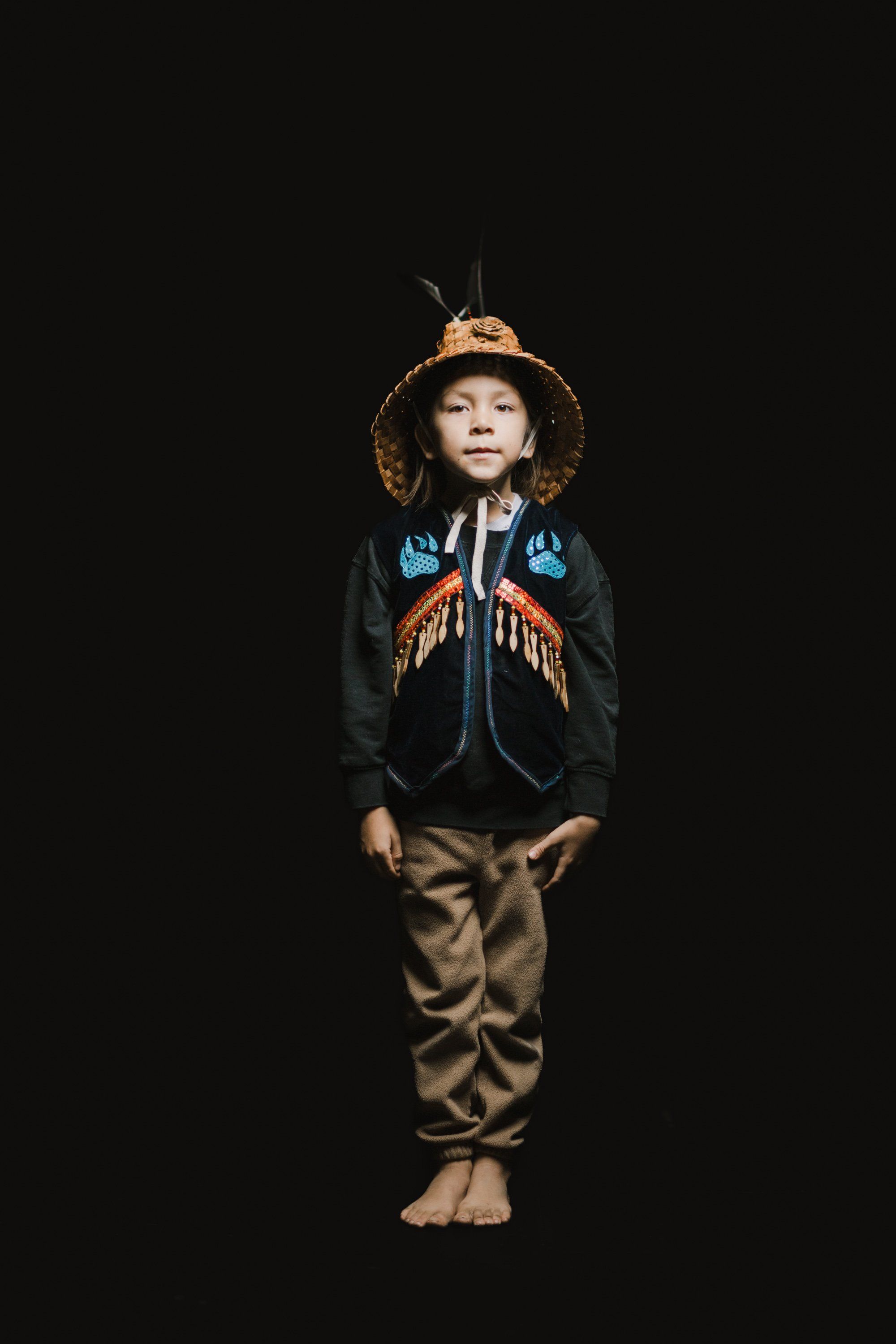 Young child in traditional Coast Salish regalia—woven hat, embroidered vest with blue and orange designs, tan pants—standing barefoot against black background