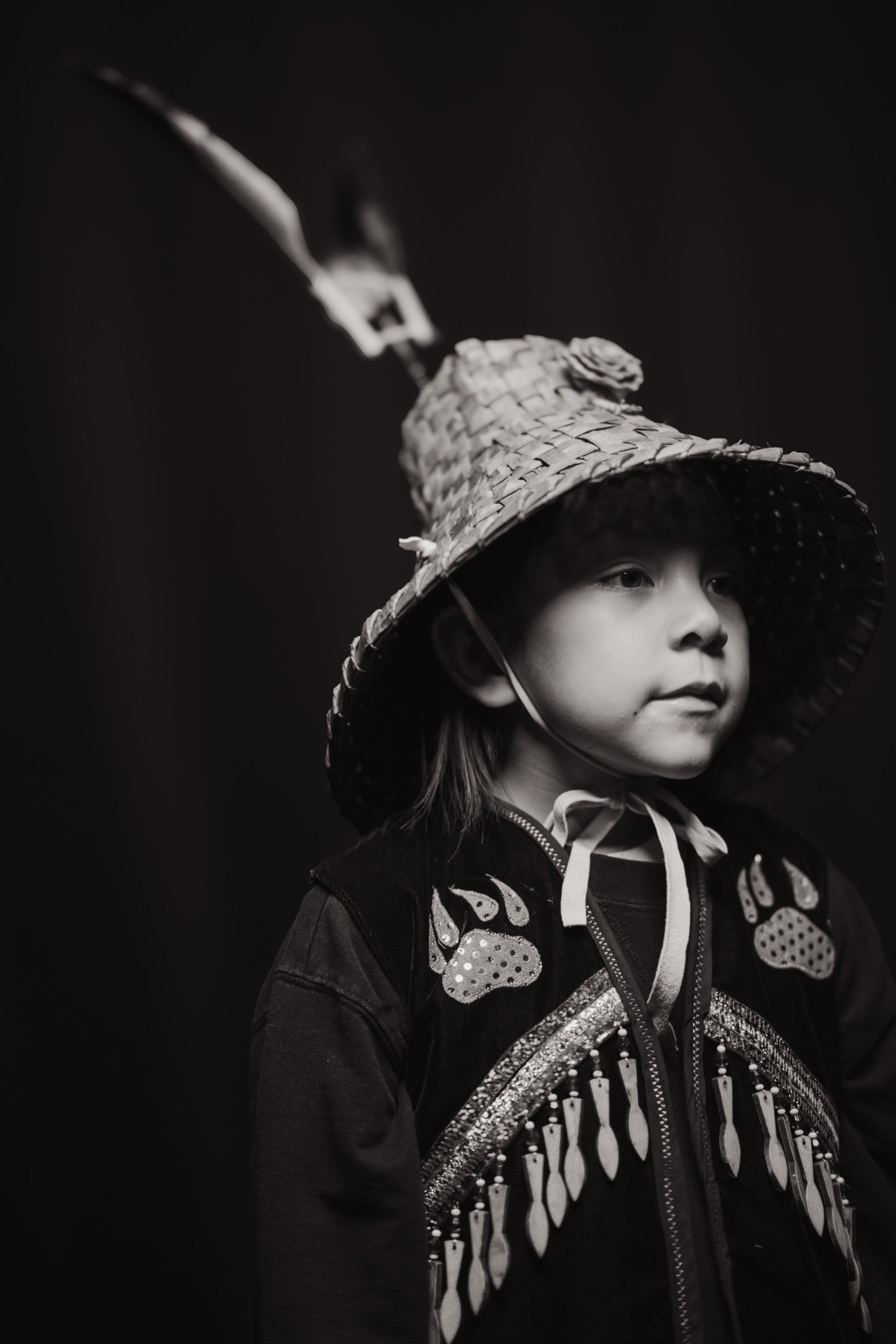 Child in traditional regalia and woven hat gazes upward against dark background, feathered ornament visible above