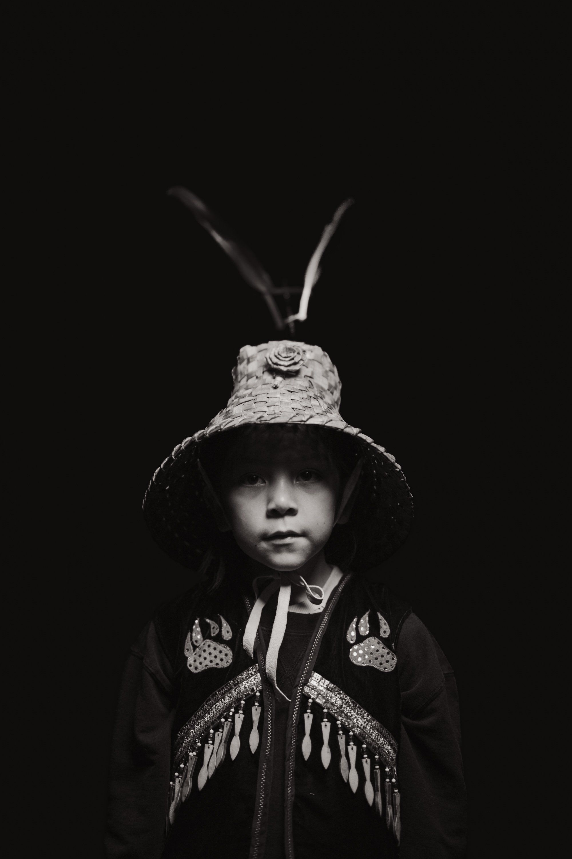 Child in traditional Coast Salish regalia with woven hat and wing ornaments, gazing downward against dark background