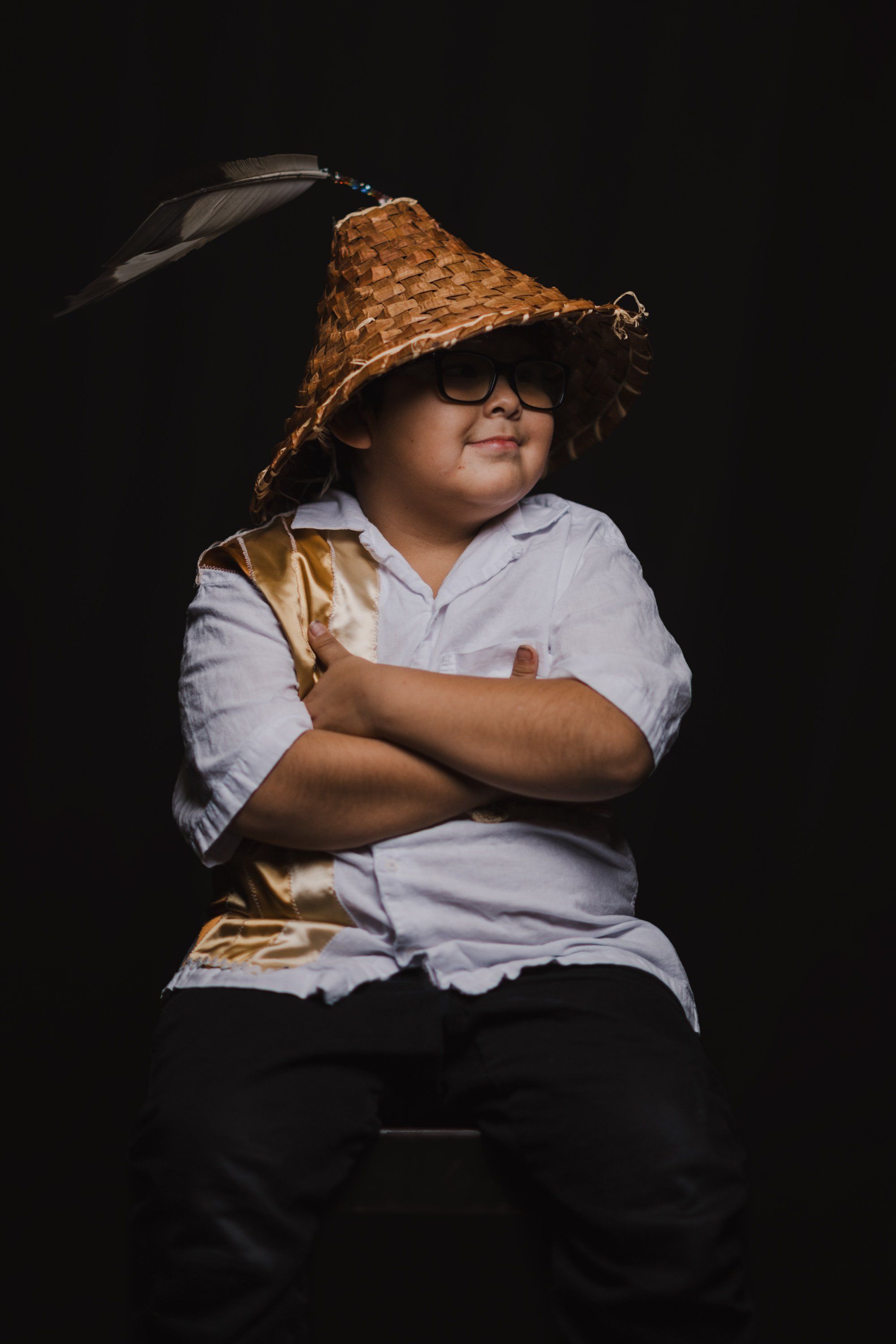 Young child wearing glasses and a woven cone hat, arms crossed, smiling warmly against a dark background with dramatic side lighting