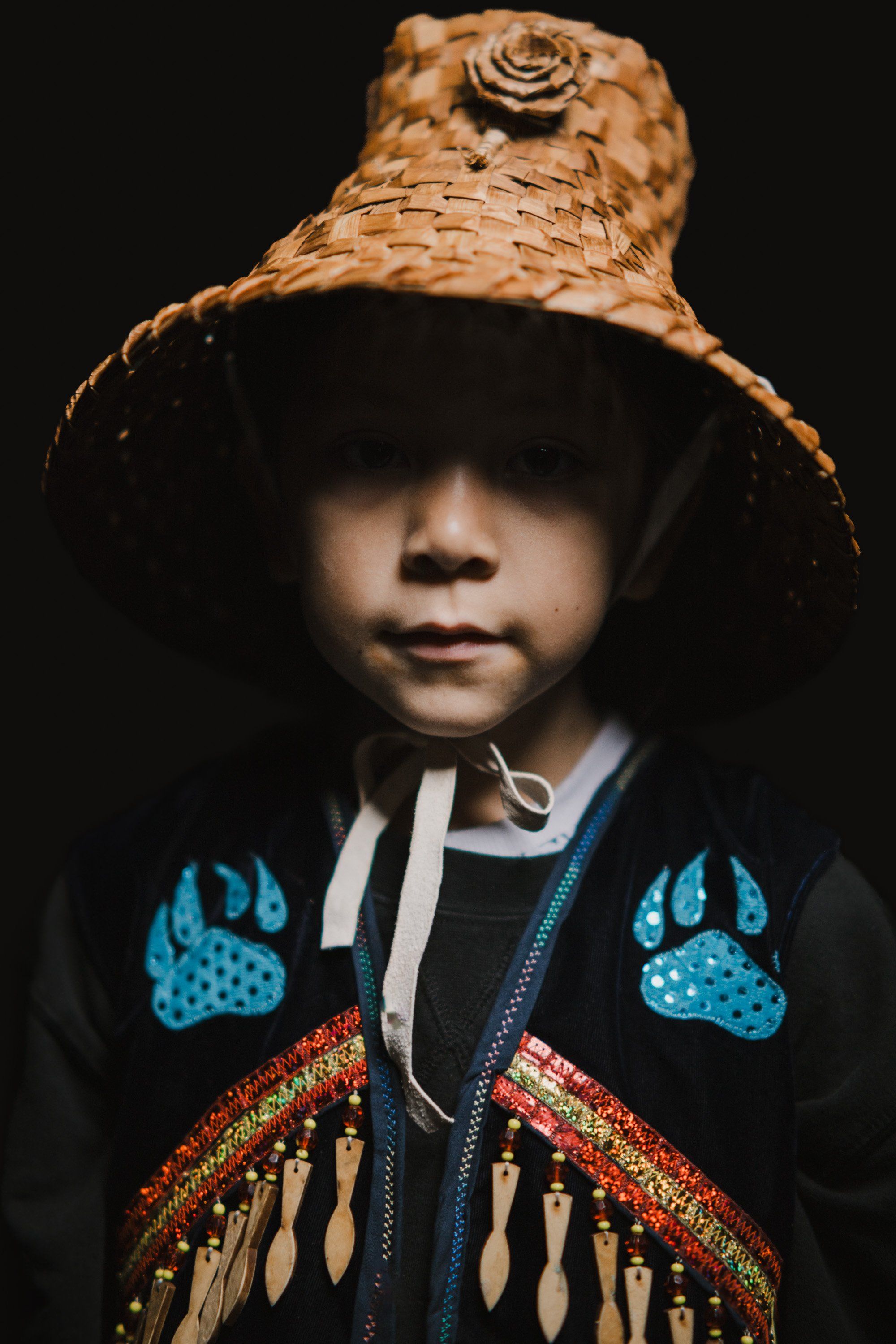 Young child in traditional Coast Salish regalia with woven hat and embroidered button blanket against dark background
