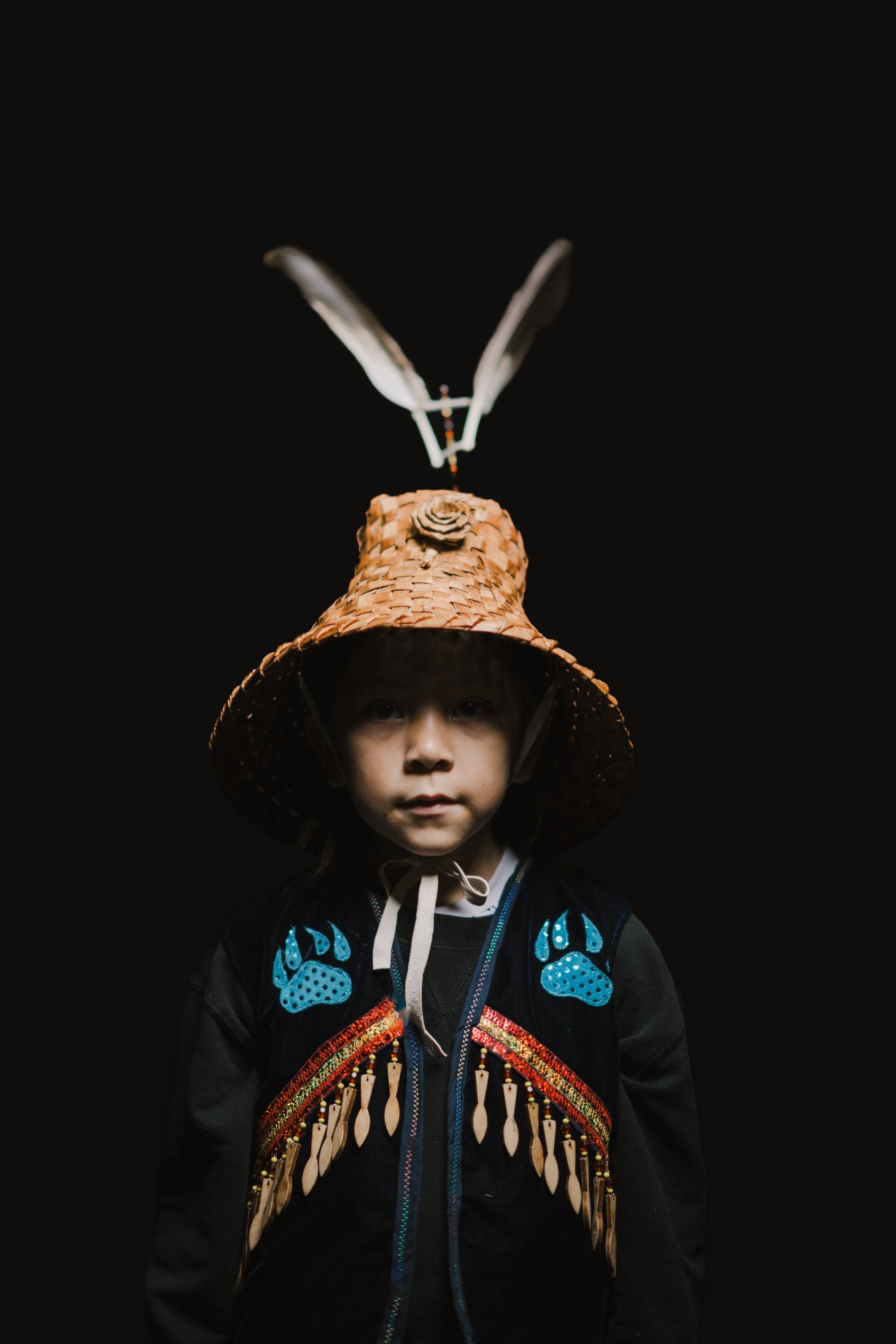 Young child wearing cork hat with feathered wings, beaded regalia against black background, embodying Coast Salish cultural tradition