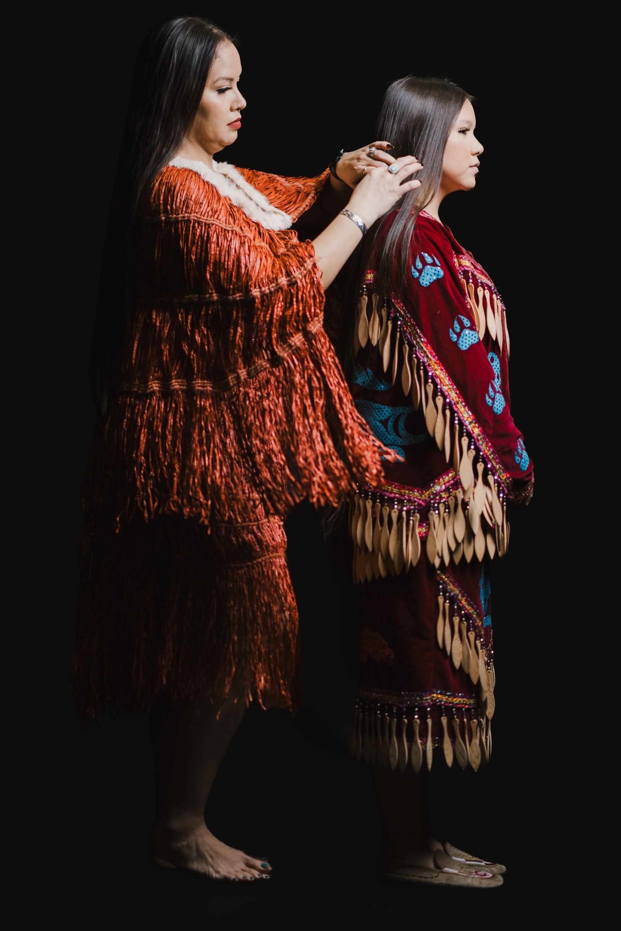 Two Coast Salish women in traditional red and burgundy button blankets with fringe stand against black, one braiding the other's hair in an intimate moment o…