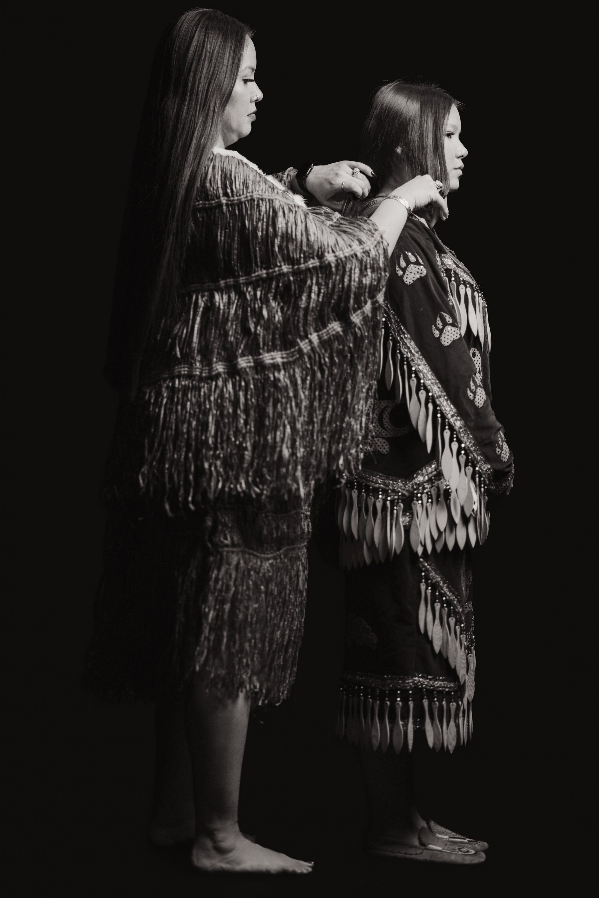 Two figures in fringed regalia stand back-to-back against black, their long dark hair flowing, captured in stark monochromatic light