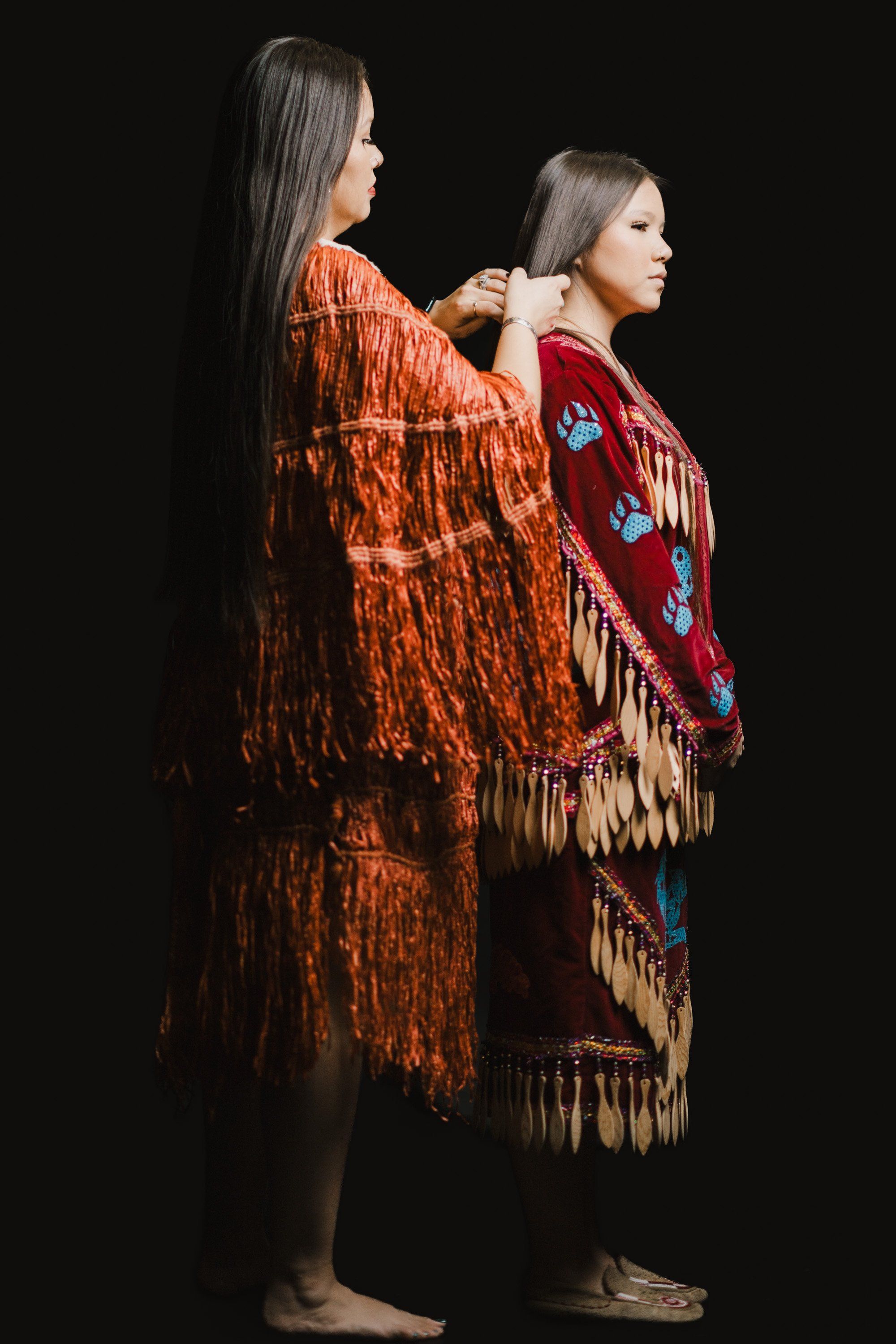 Two young people in ornate fringed regalia stand against black, one in rust orange and one in deep red with blue designs, facing right in profile