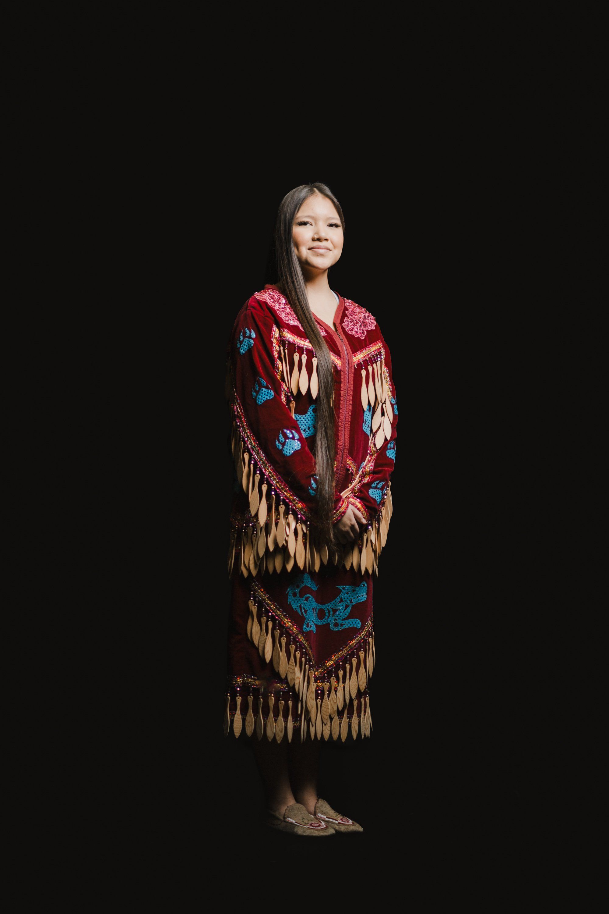 Woman in red and gold fringed Coast Salish regalia smiles against black background, embodying traditional dance attire