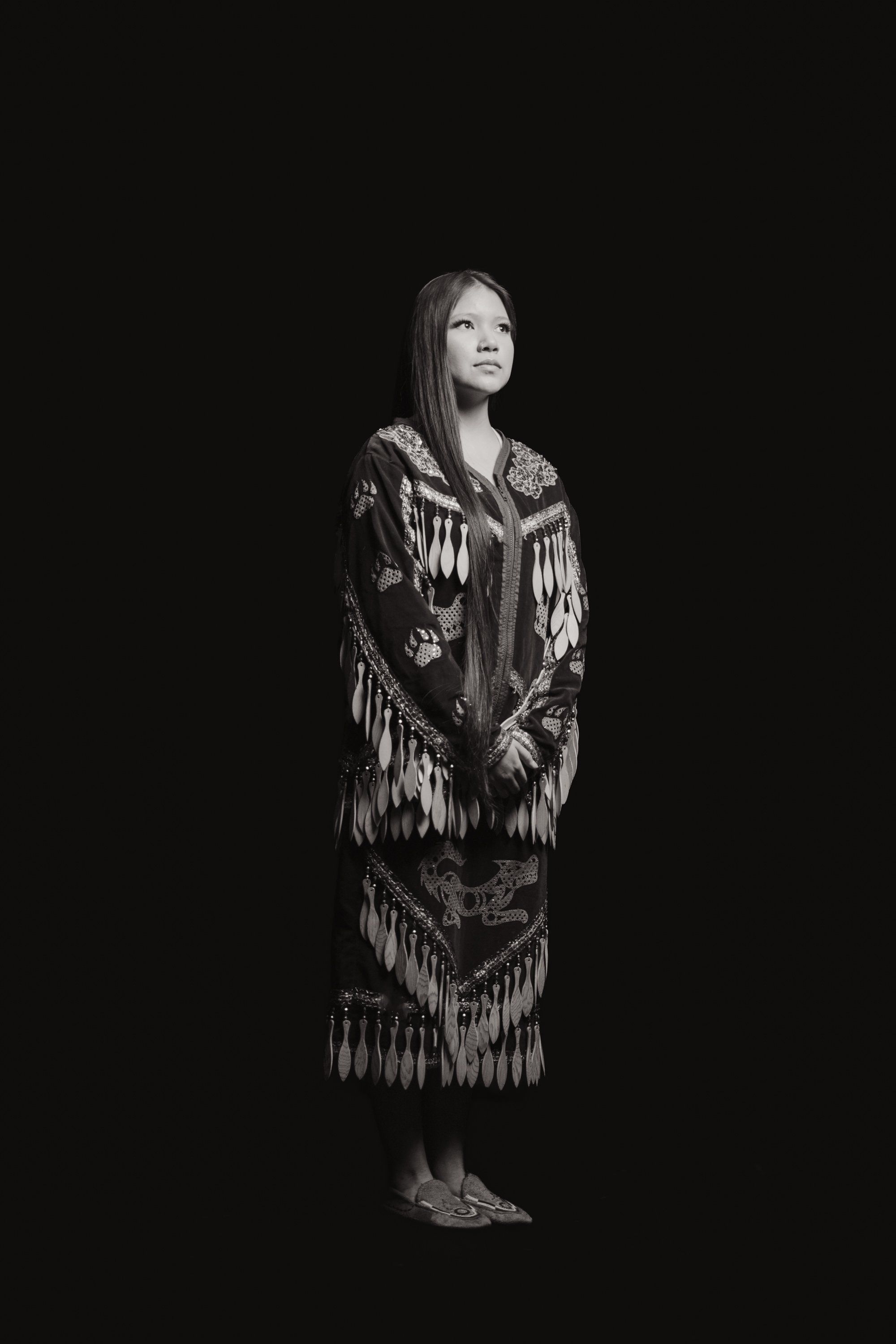 Woman in traditional fringed regalia stands against black background, gazing upward in contemplative pose