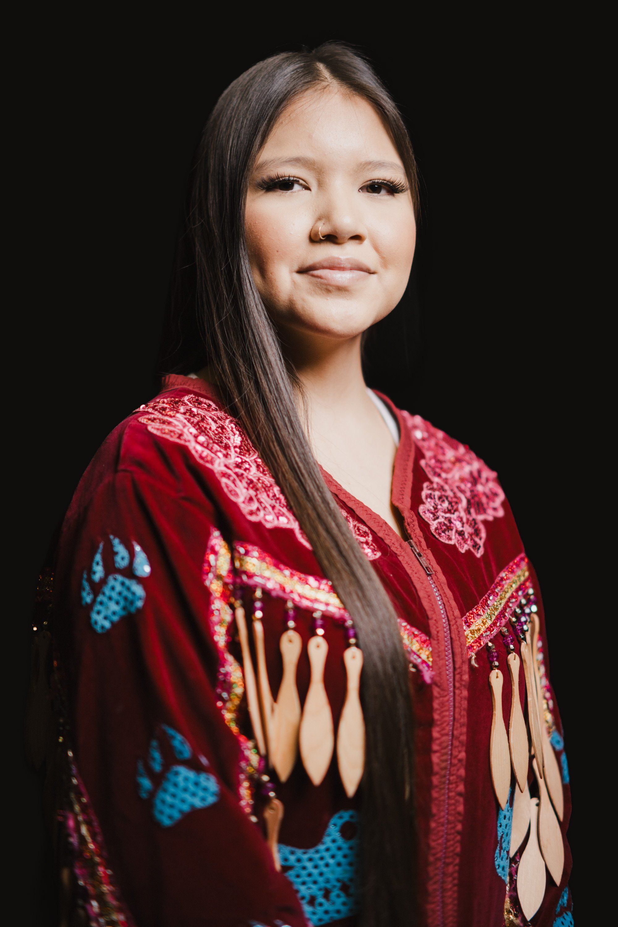 Young woman in burgundy Coast Salish regalia with blue and cream geometric patterns and fringe, long dark hair, against black background