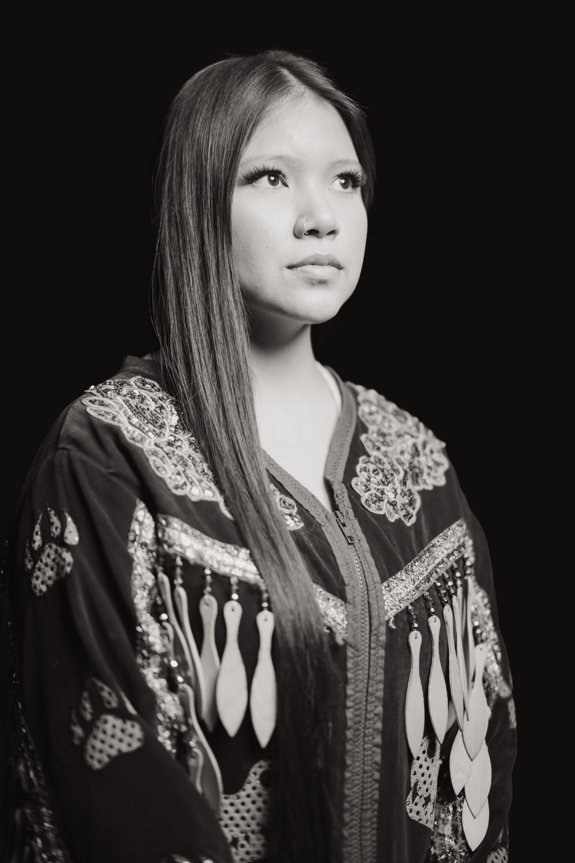 Young person in traditional Coast Salish regalia with geometric patterns, long dark hair, gazing upward against black background