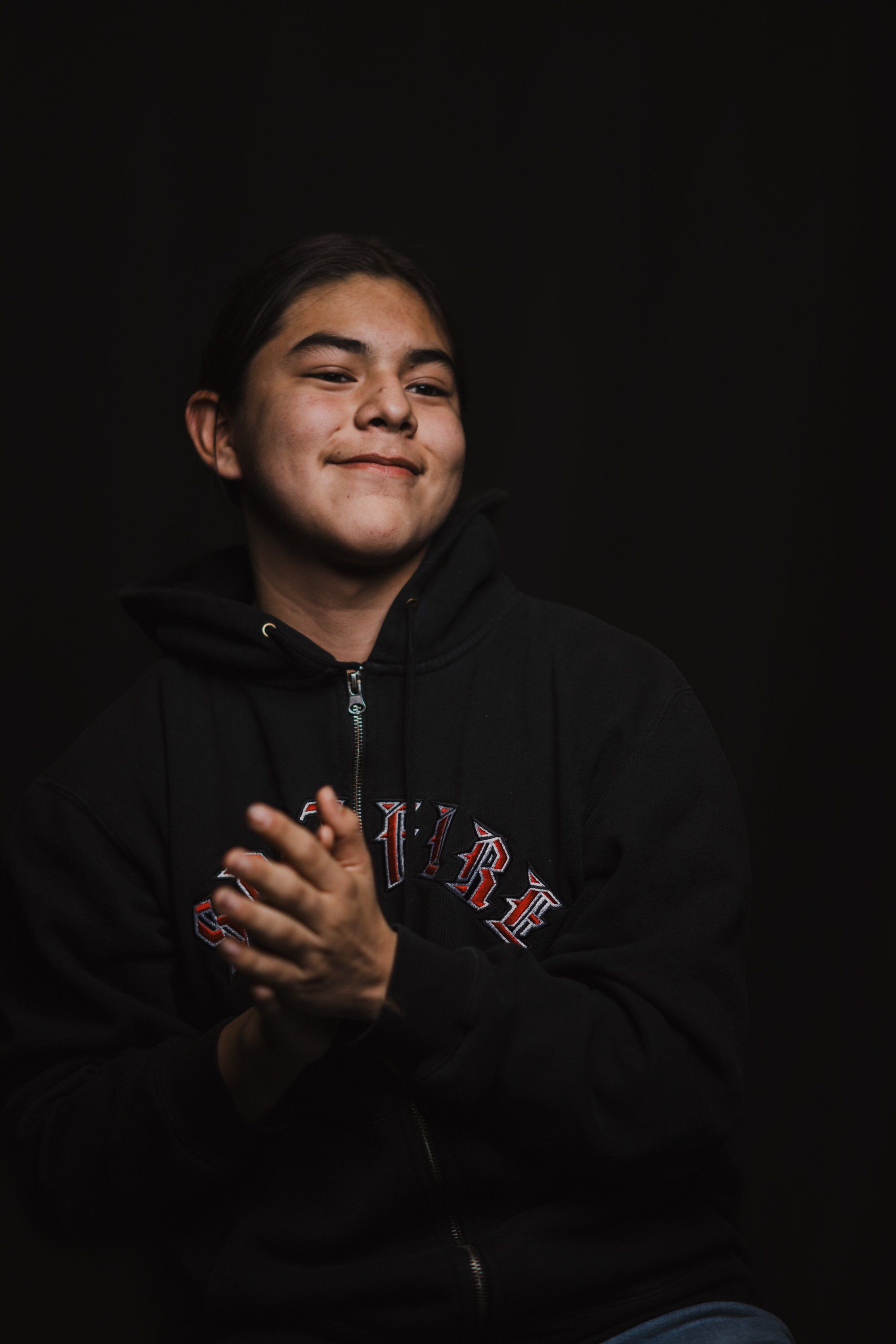 Young person in black zip hoodie with red text, hands clasped at chest, gazing upward against dark background with warm sidelighting