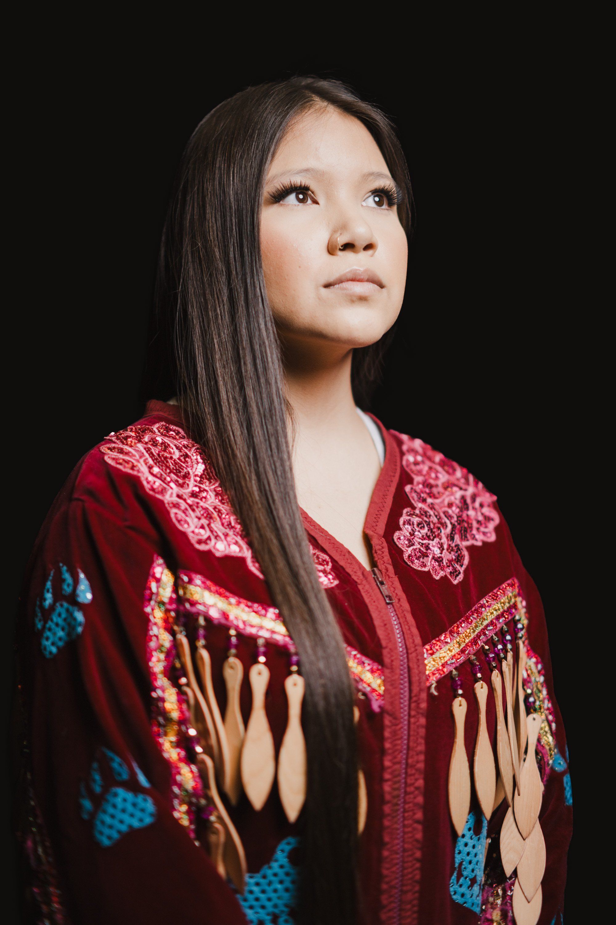 Young woman in ornate red and blue button blanket with fringe against black background, gazing upward with contemplative expression