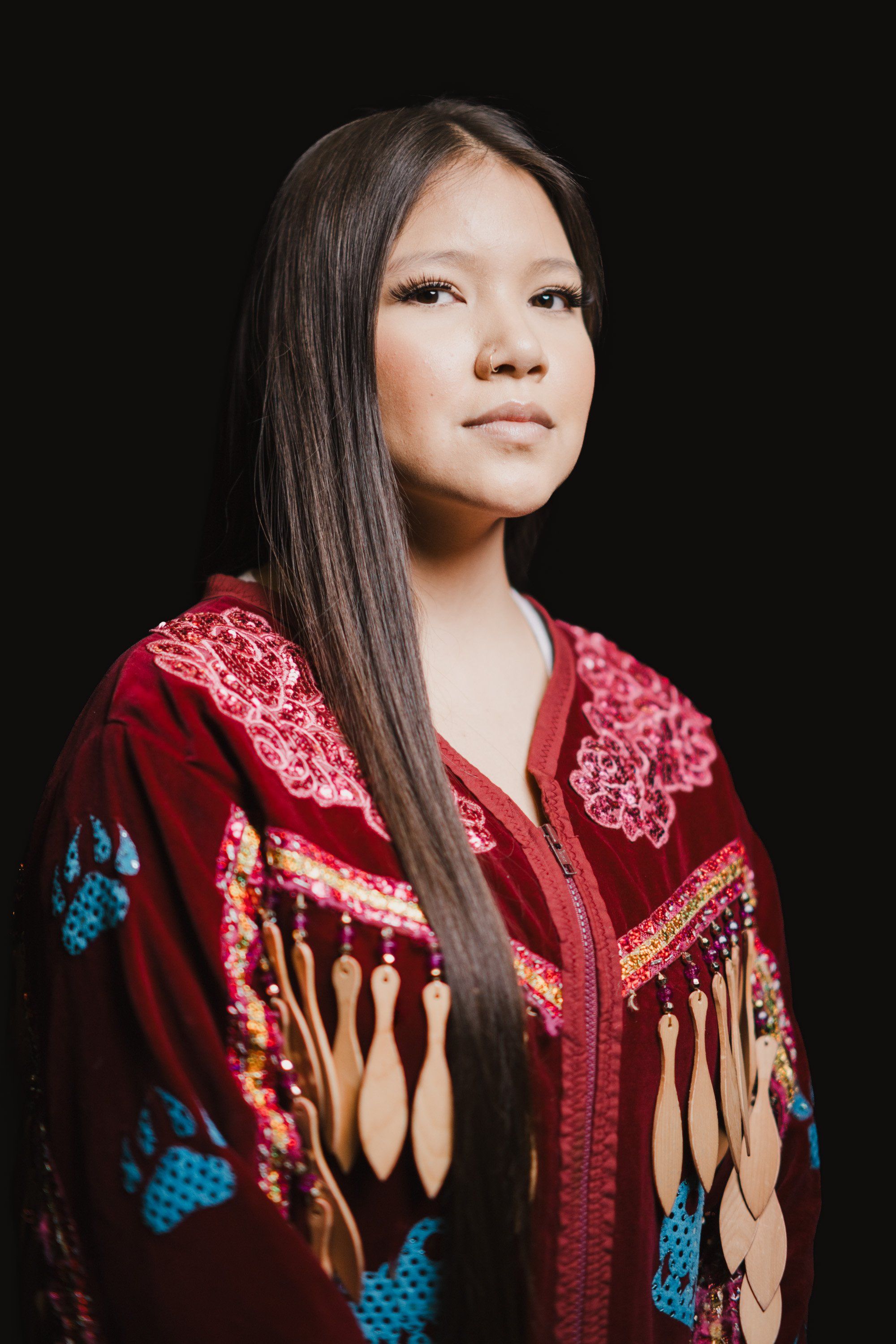 Young woman in burgundy beaded traditional Coast Salish regalia with fringe and blue accents, long dark hair, against black background