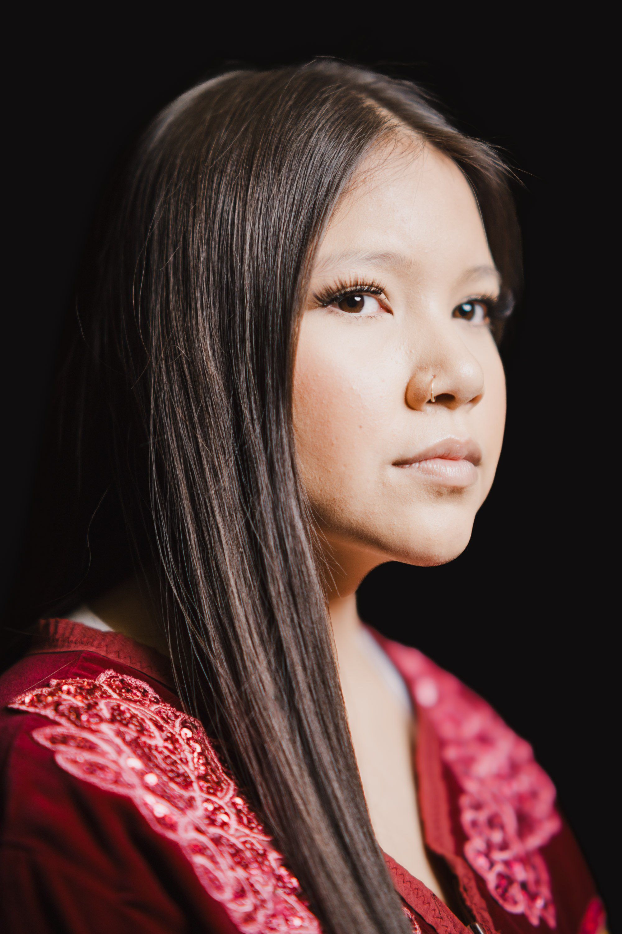 Woman in crimson traditional regalia with intricate beadwork, long dark hair, gazing thoughtfully against black background