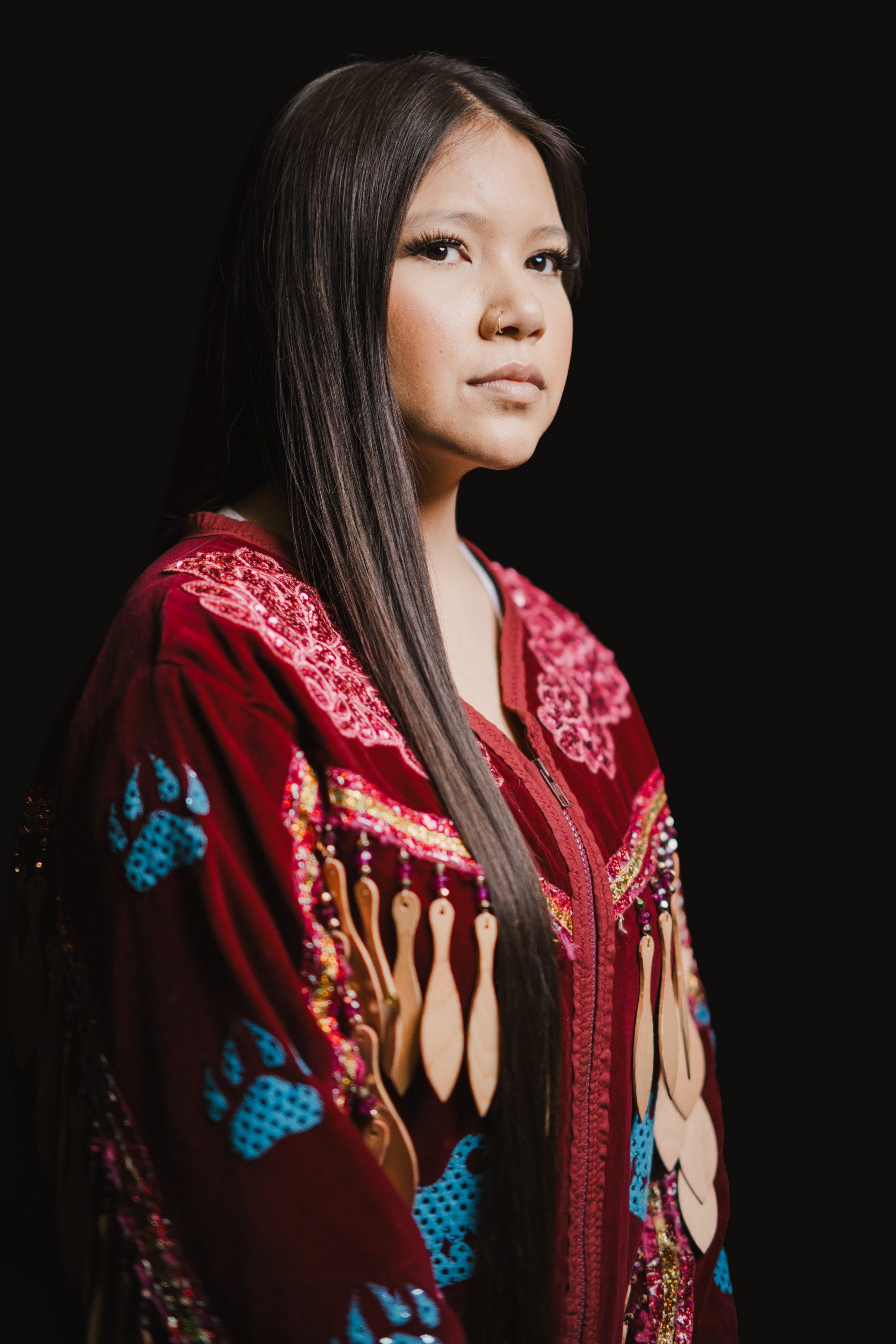 Woman in deep red embroidered Coast Salish regalia with geometric patterns, long dark hair, gazing thoughtfully against black background