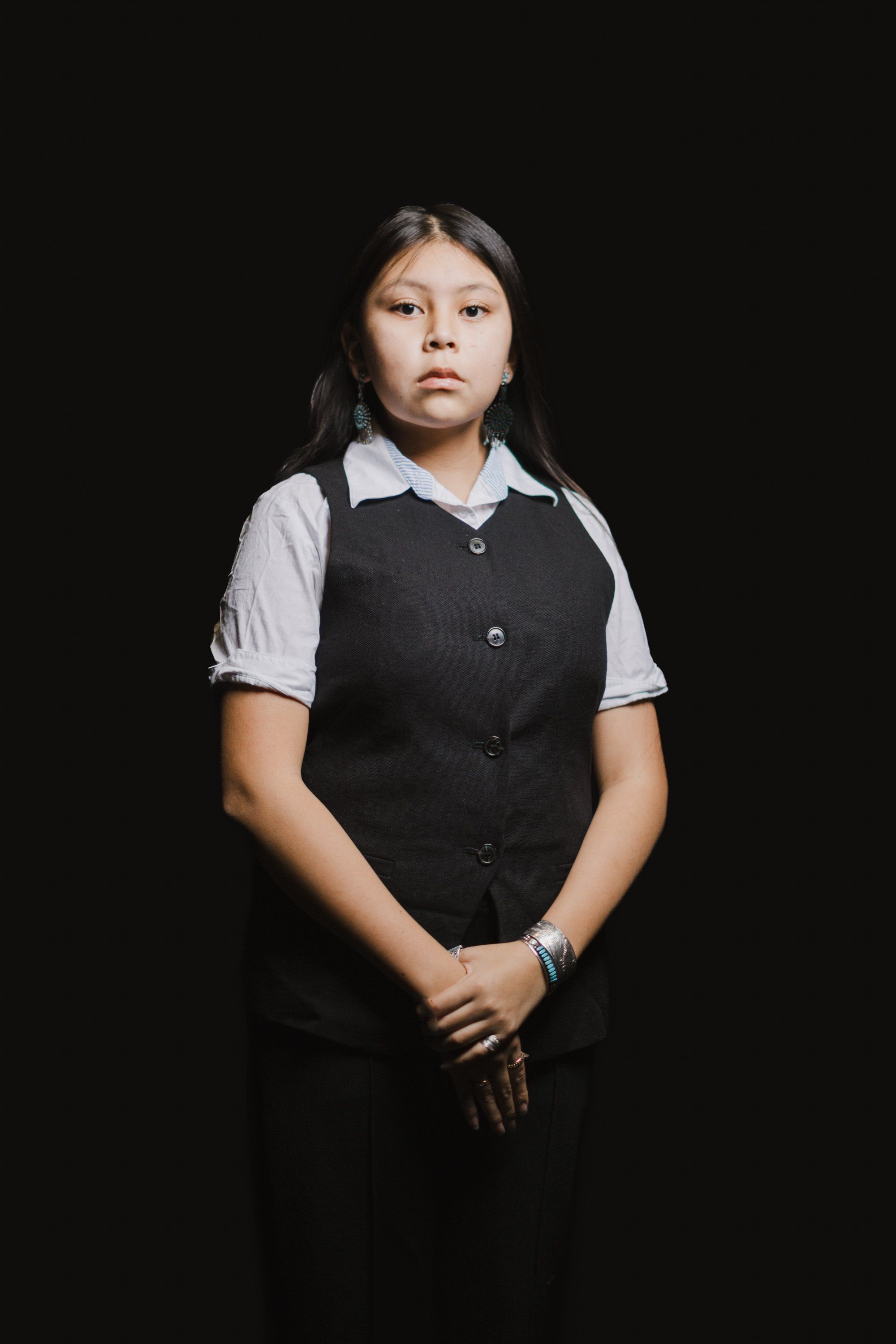Young person in black vest and white collared shirt stands against dark background, hands clasped, direct gaze