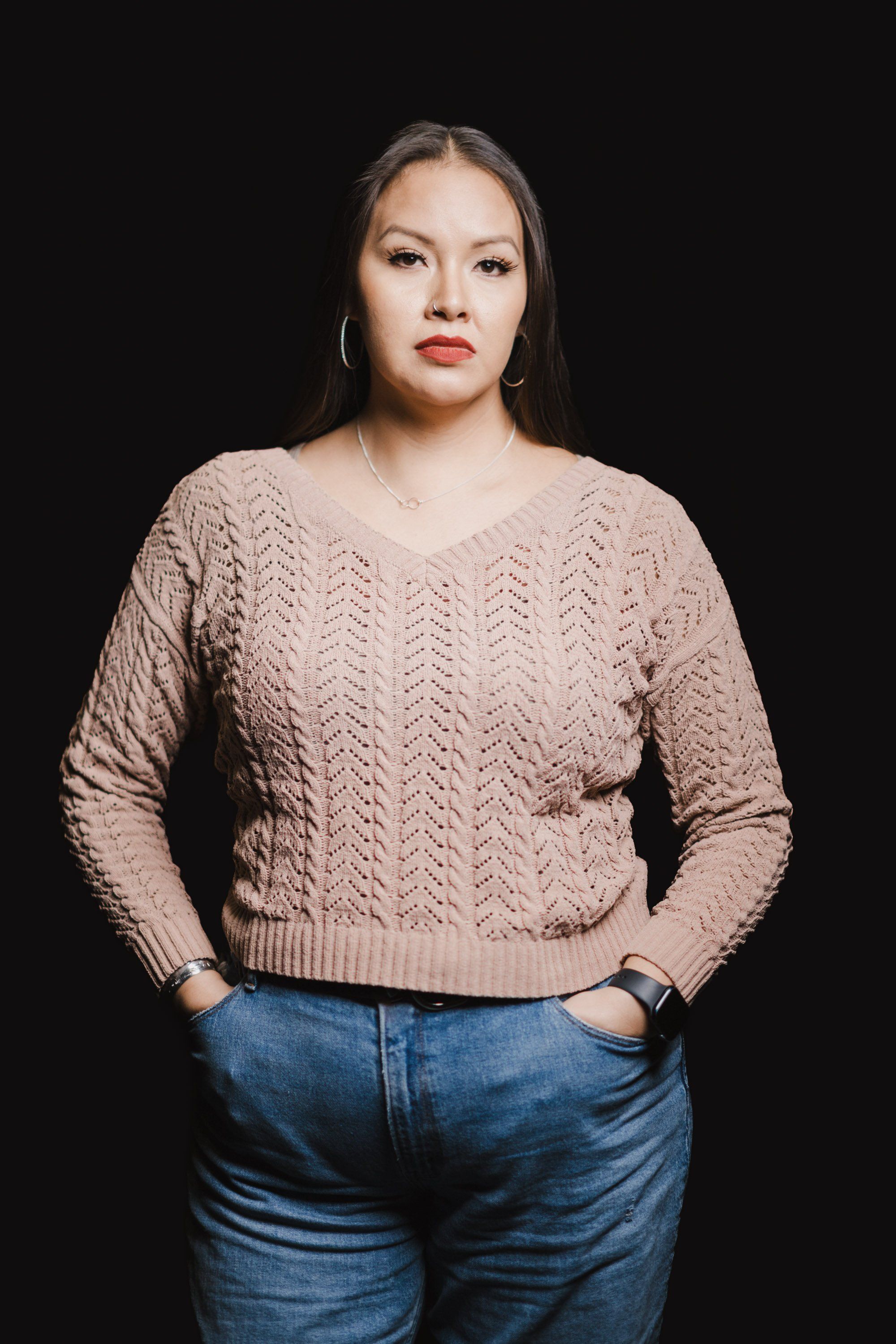 Woman in taupe cable-knit sweater and blue jeans poses against black background, hands in pockets, direct gaze