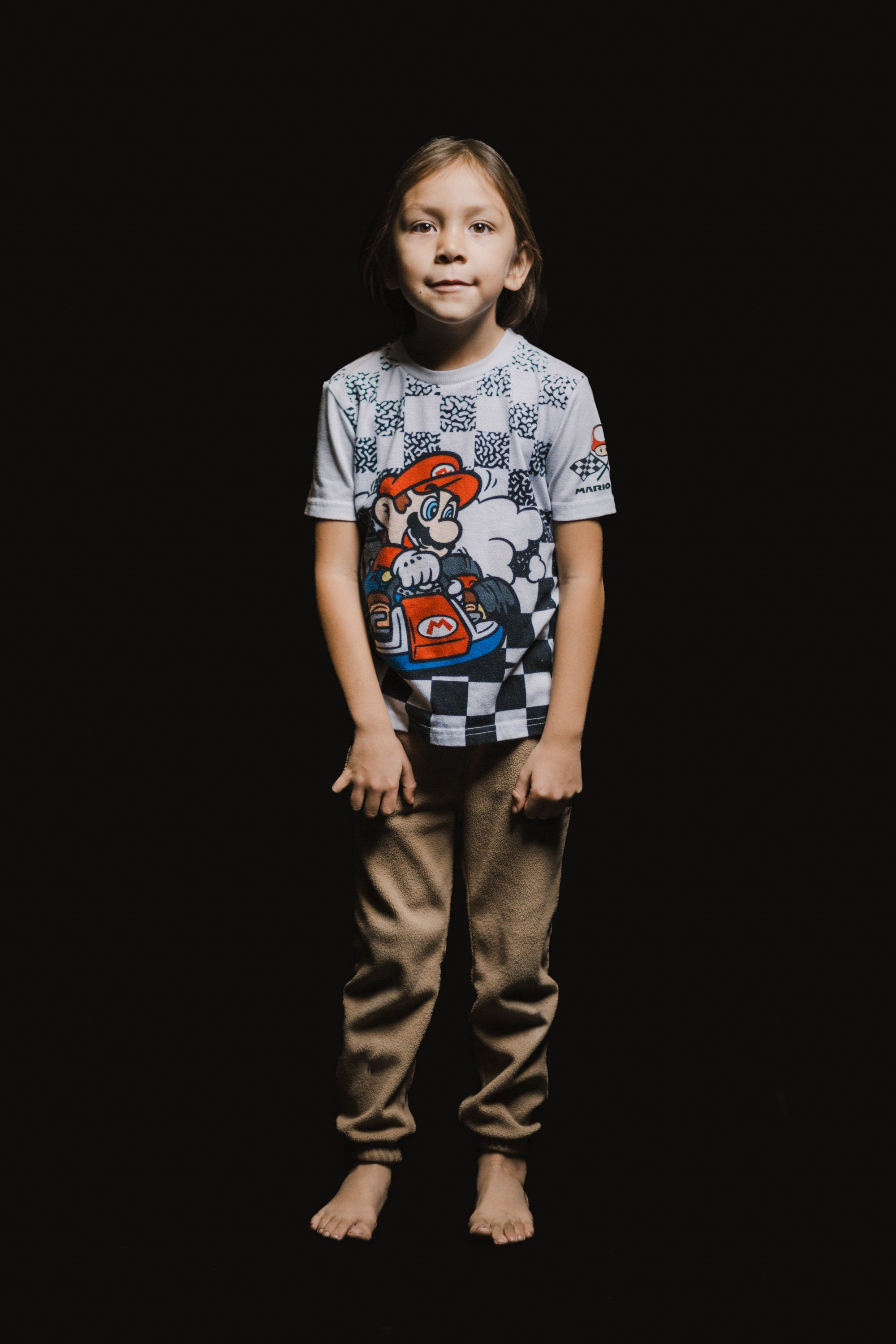 Young child in graphic t-shirt and tan pants stands barefoot against black background, gazing upward with contemplative expression