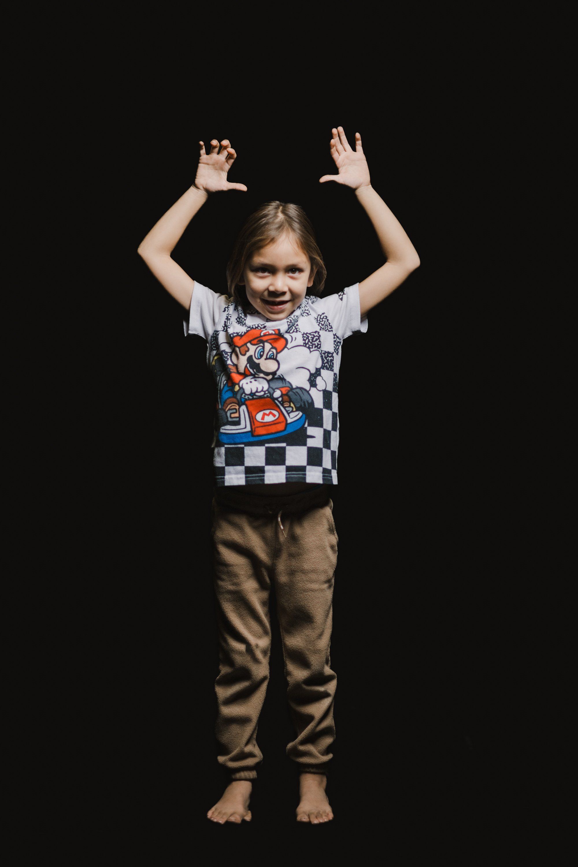 Young child in checkered shirt with arms raised joyfully against black background, barefoot and smiling