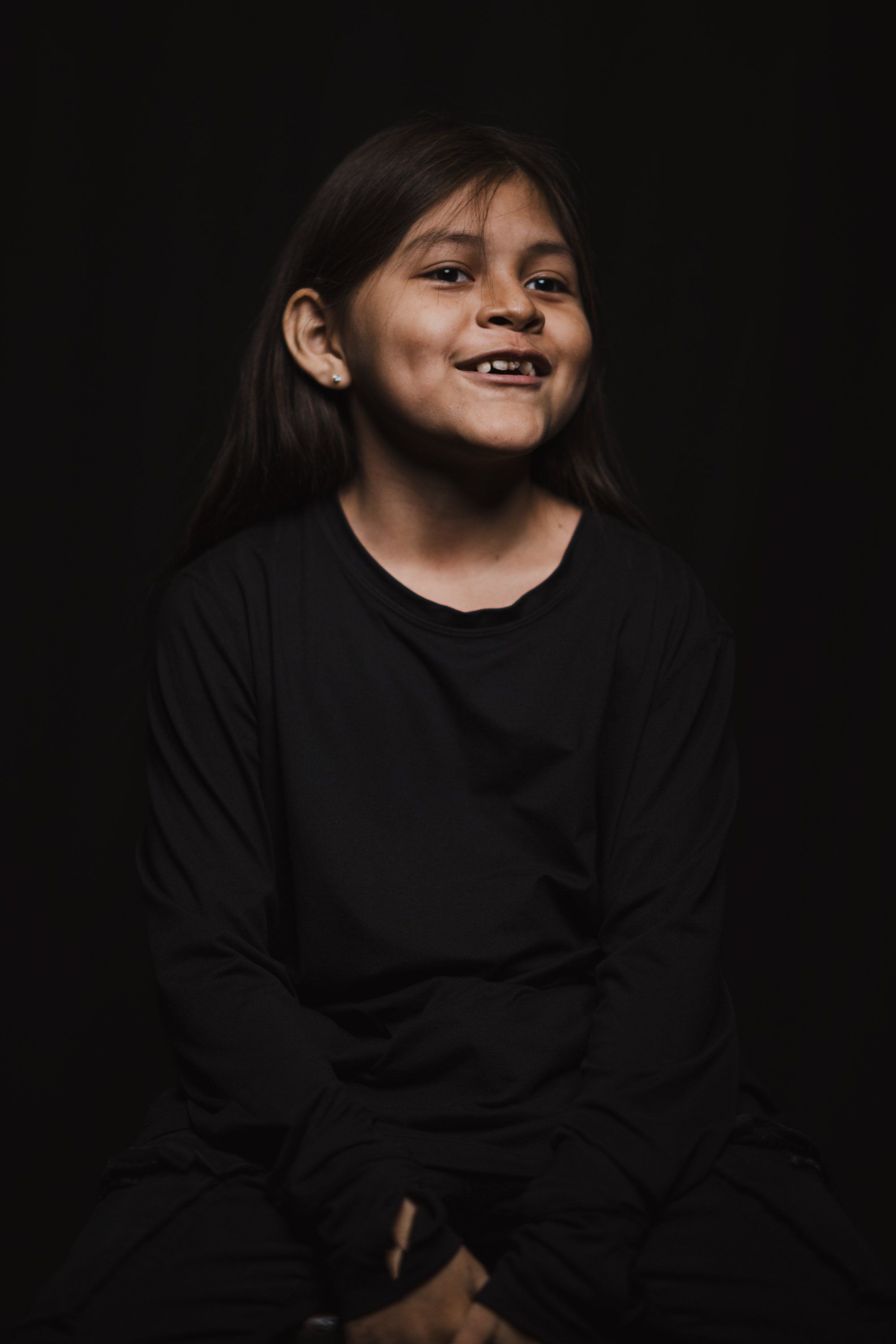 Young person in black long-sleeve shirt smiling against dark background, warm studio lighting illuminating their face