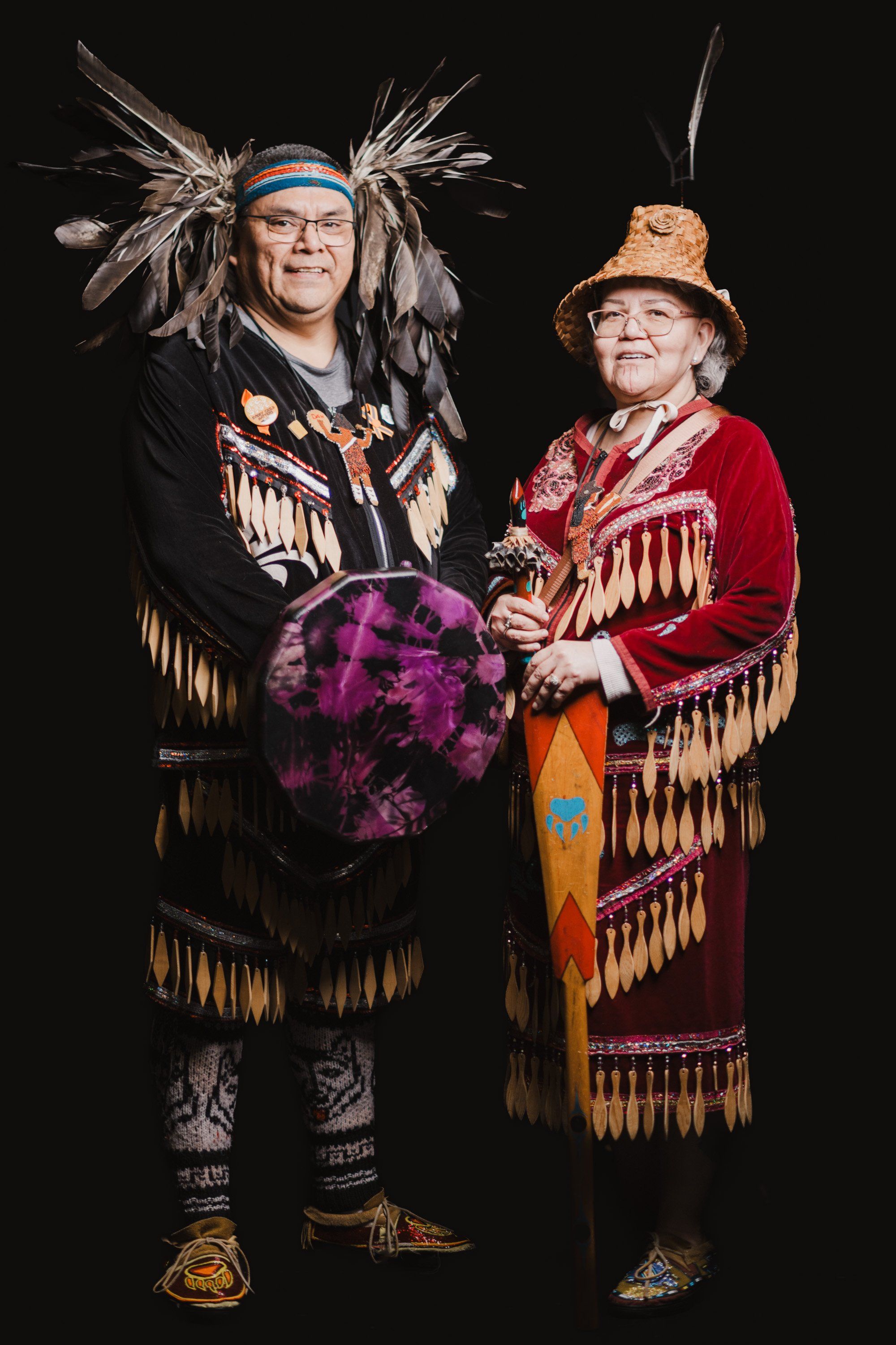 Two Coast Salish dancers in traditional regalia with fringe and feathered headdresses pose against black, holding a purple drum
