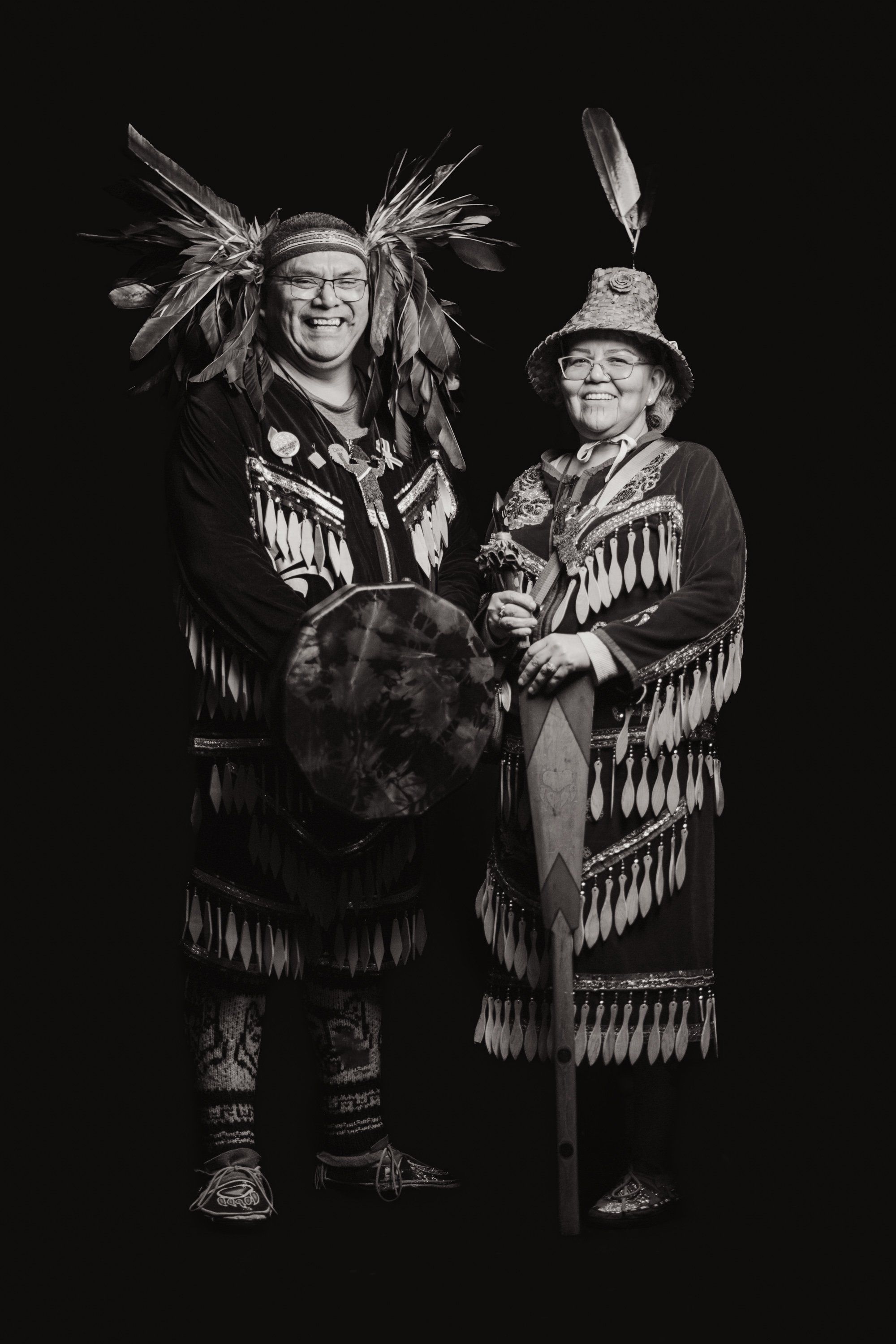 Two people in traditional Coast Salish regalia with fringed button blankets and feathered headdresses stand together against black, holding a drum between them
